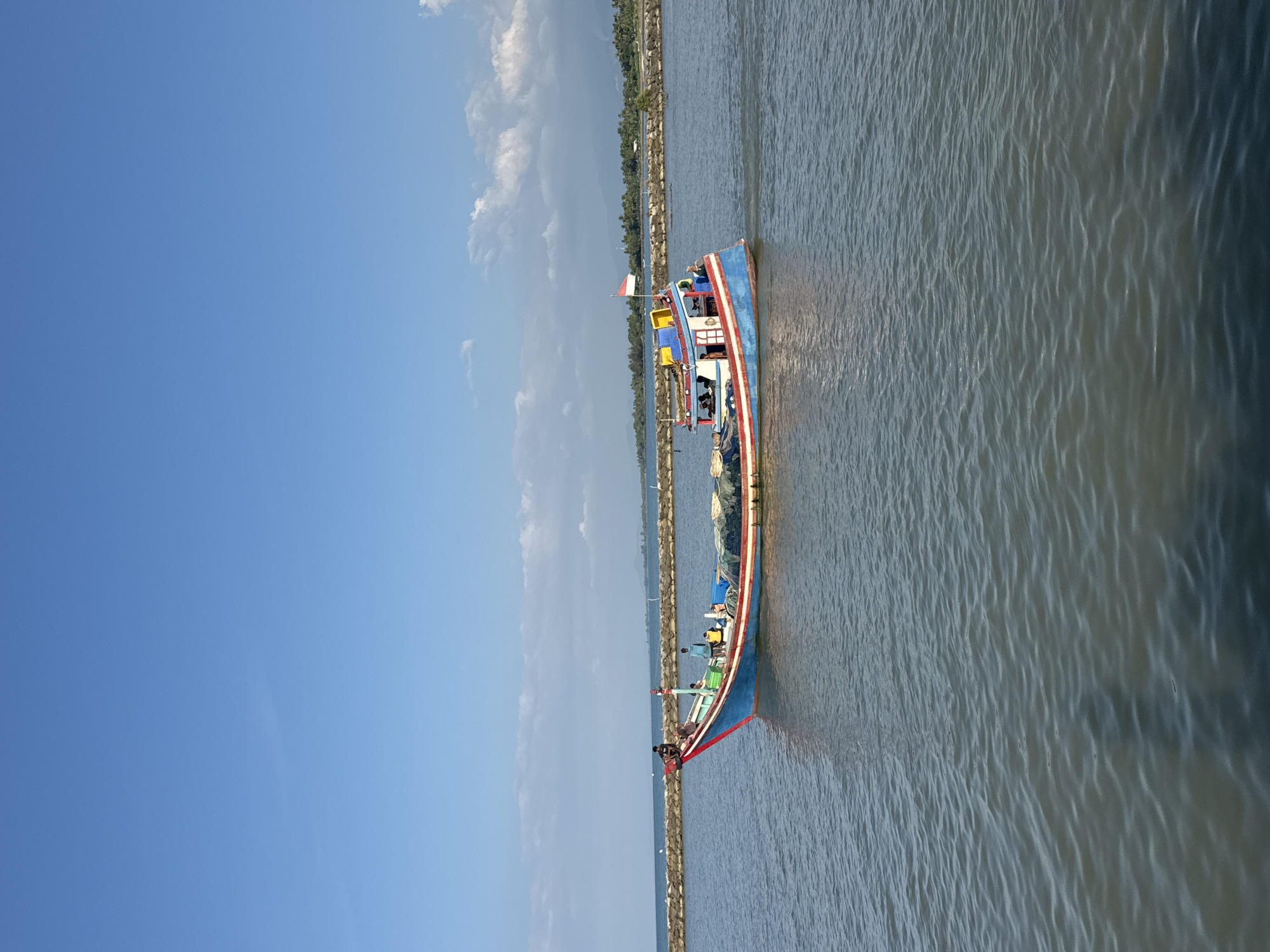 A medium-sized blue and red fishing boat sails on a body of water under a bright blue sky with scattered white clouds. The boat, which appears to be a traditional Indonesian vessel, has a brightly colored cabin structure with yellow and red accents. Several people are visible on board, some standing and some sitting. A red and white Indonesian flag is flying from a pole on the boat's cabin. In the background, a stone breakwater runs along the coastline, with lush green vegetation and palm trees visible beyond it. The water is calm with gentle ripples. The overall scene depicts a sunny day at sea near a coastal area.