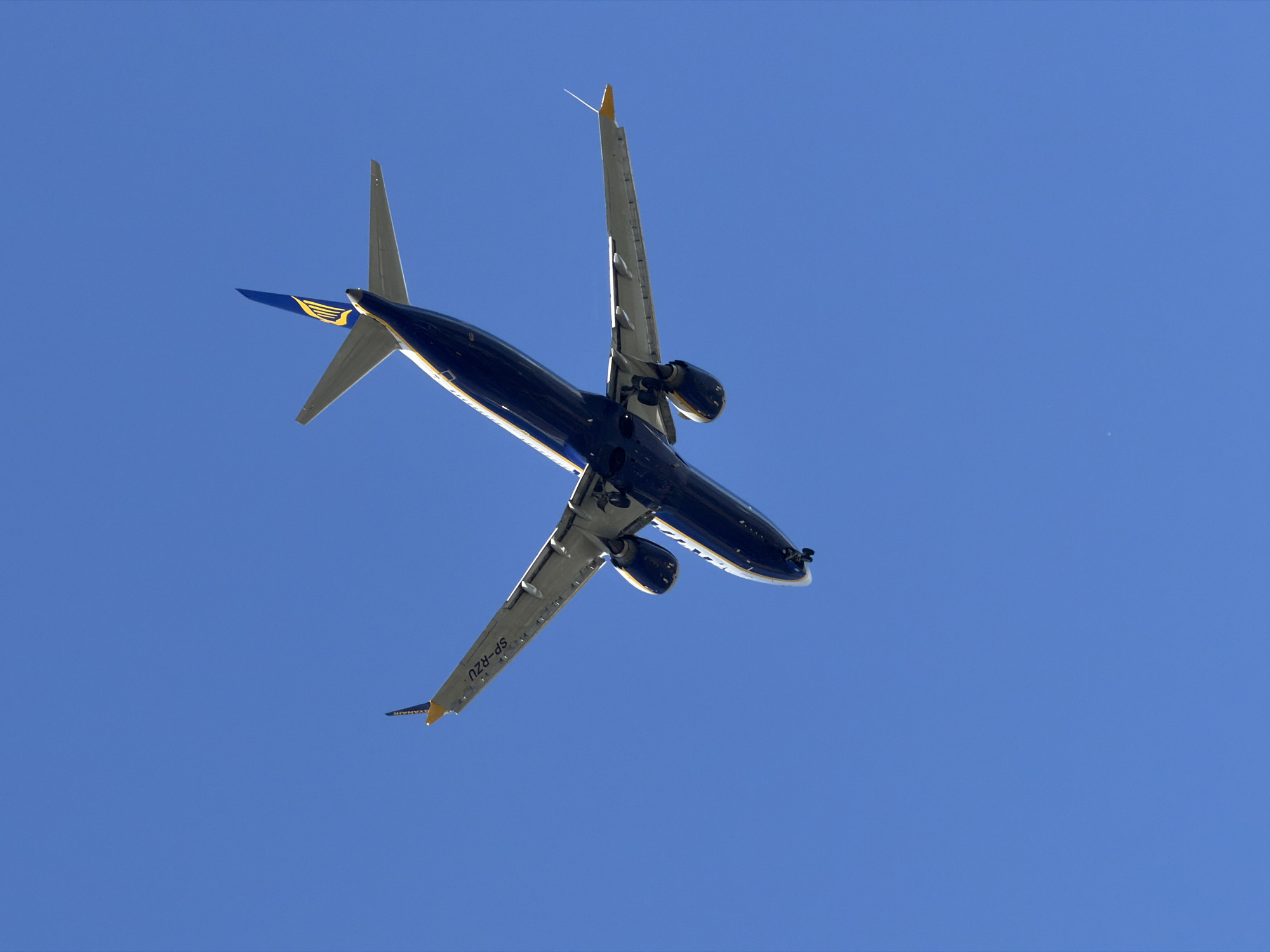 An underside view of a dark blue and yellow Ryanair airplane is captured against a clear, bright blue sky. The aircraft is angled upwards, with its tail and a portion of its wings visible. The registration number "SP-RZU" is visible on the wing. The plane appears to be in flight, possibly ascending or descending. The image does not provide any specific landmarks or context that would indicate its location within the city of Burgas, Bulgaria.