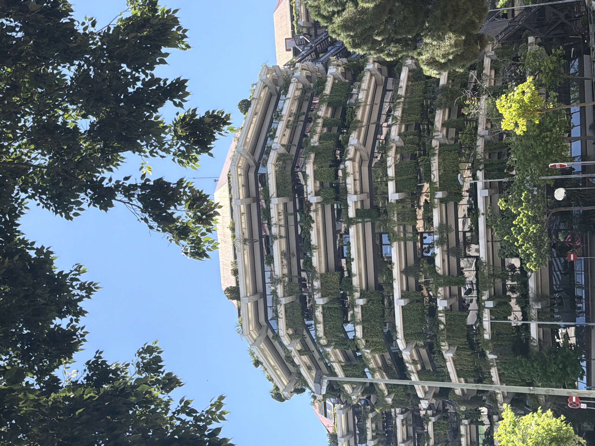 The image shows the residential building on Carrer de Praga, Barcelona, Spain, with its distinctive vegetation. The building has a stepped facade with plants growing on each level. The building is partially obscured by trees in the foreground and background. The sky is visible through the trees. A streetlight is on the right side of the image.
