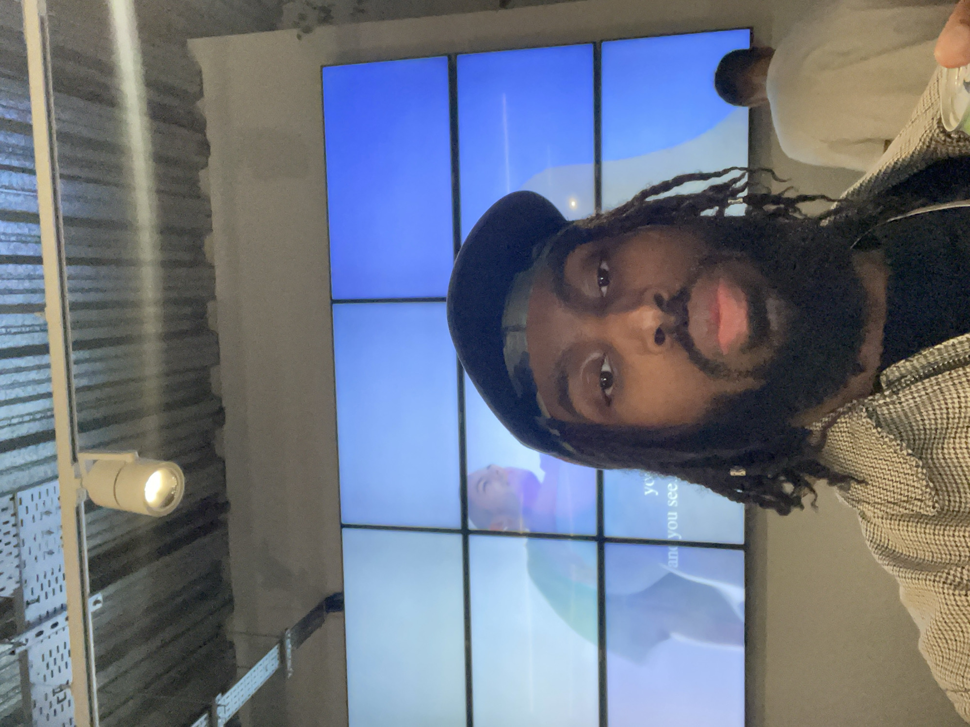 A selfie captures a man with dark skin, dreadlocks, and a dark cap/beanie, looking directly at the camera with a neutral to slightly serious expression. He has a beard and mustache. He is wearing a black shirt and a patterned jacket (houndstooth or similar plaid) over it.
In the background, a large video wall is prominent, made up of multiple rectangular screens displaying abstract blue and light-colored graphics. Partially visible text on these screens reads "you" and "and you see." Another person, wearing a light-colored top, is partially visible in the upper right portion of the image, possibly reflected on the screen or standing behind the main subject.
To the left, a white, cylindrical track light fixture with an illuminated bulb is mounted on a vertical pole, providing direct lighting. The wall behind this light fixture exhibits an industrial aesthetic, with a textured surface that appears to be corrugated or segmented, possibly exposed concrete or metal paneling.
The setting appears to be an indoor, modern space, potentially an exhibition hall, gallery, or contemporary retail environment in London, United Kingdom. The bright screens and focused lighting suggest an active, designed space.