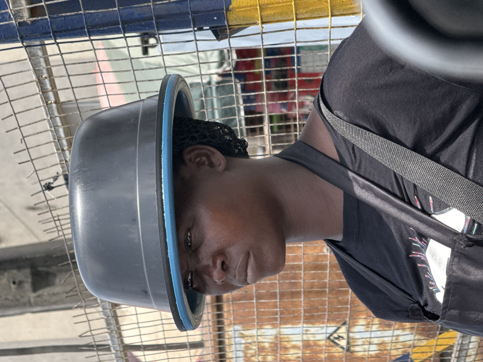 A woman in Lagos, Nigeria, wears a large gray plastic bucket on her head. The bucket has a blue rim. She is wearing a black t-shirt and a gray strap is slung over her shoulder. She is standing in front of a chain link fence. Behind the fence there are multiple colors, including yellow and red. The ground is made of concrete.
