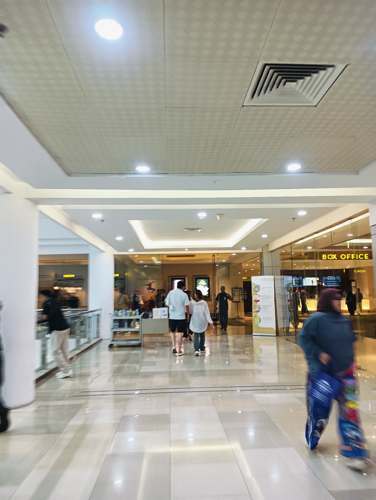 The image captures a well-lit indoor shopping mall corridor in Batam, Indonesia. The ceiling features recessed lighting and a square air vent. In the background, a "BOX OFFICE" sign is visible on a glass-fronted establishment, suggesting a cinema complex. Several individuals are present, some walking and others partially visible. A person in the foreground is blurred by motion and wears a blue jacket and colorful patterned pants, carrying a blue bag. Further into the corridor, two individuals in casual attire are walking away from the camera, and other people are seen near shop entrances. The flooring is polished and reflects the ambient light. The overall environment appears clean and modern, typical of a commercial establishment.