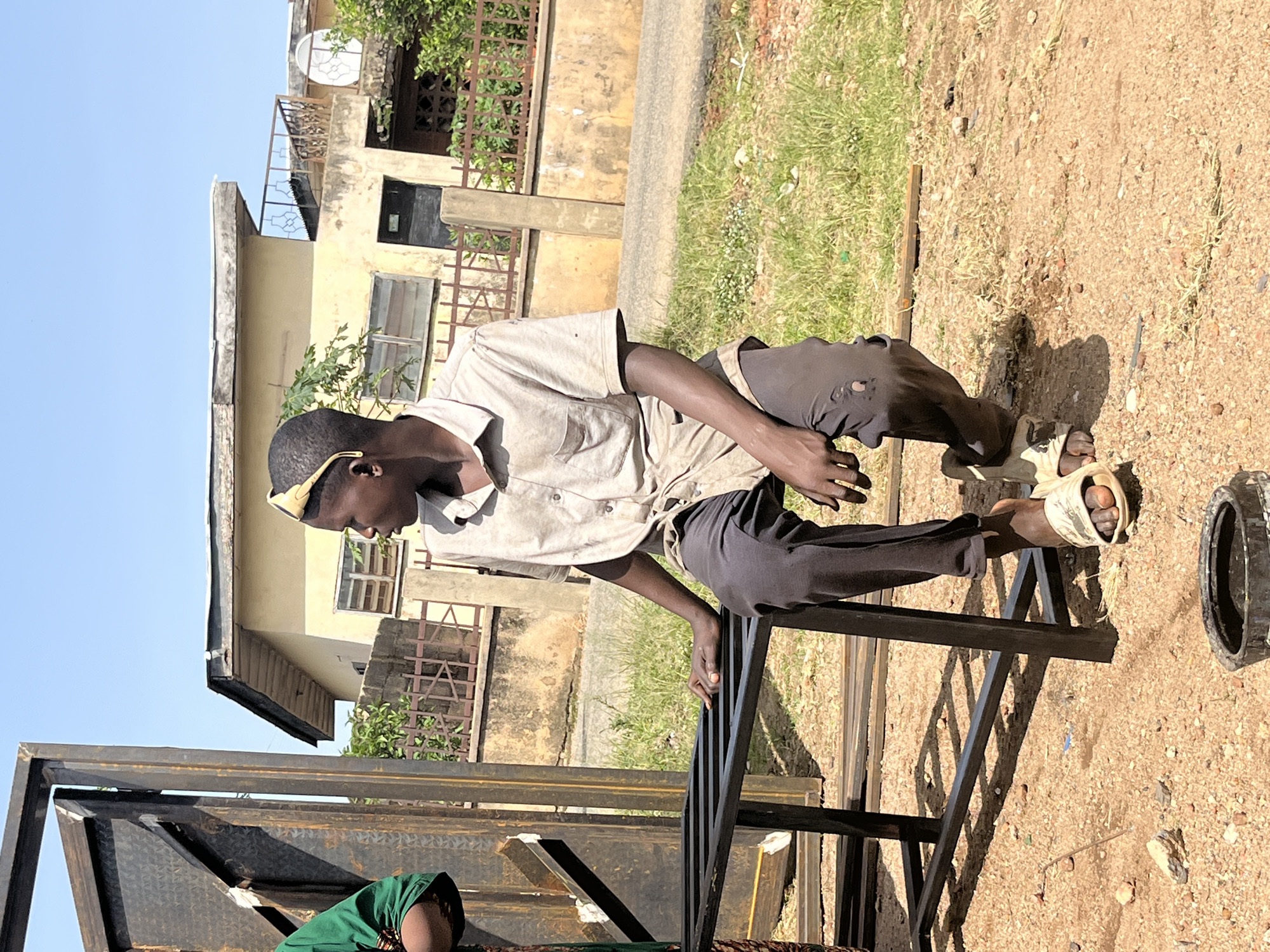 A young African male is sitting on a metal bench outdoors. He is wearing a light-colored short-sleeved collared shirt and dark pants. He has yellow sunglasses on his forehead. The man is looking to his right, with his left hand resting on the bench beside him. The background shows a light-colored building, a fence, and some trees. The ground is covered in dirt and grass. The image appears to be taken in an urban or semi-urban environment. The person is possibly working or taking a break from work. The scene is brightly lit by sunlight. The specific location is within the city of Jos, Nigeria.