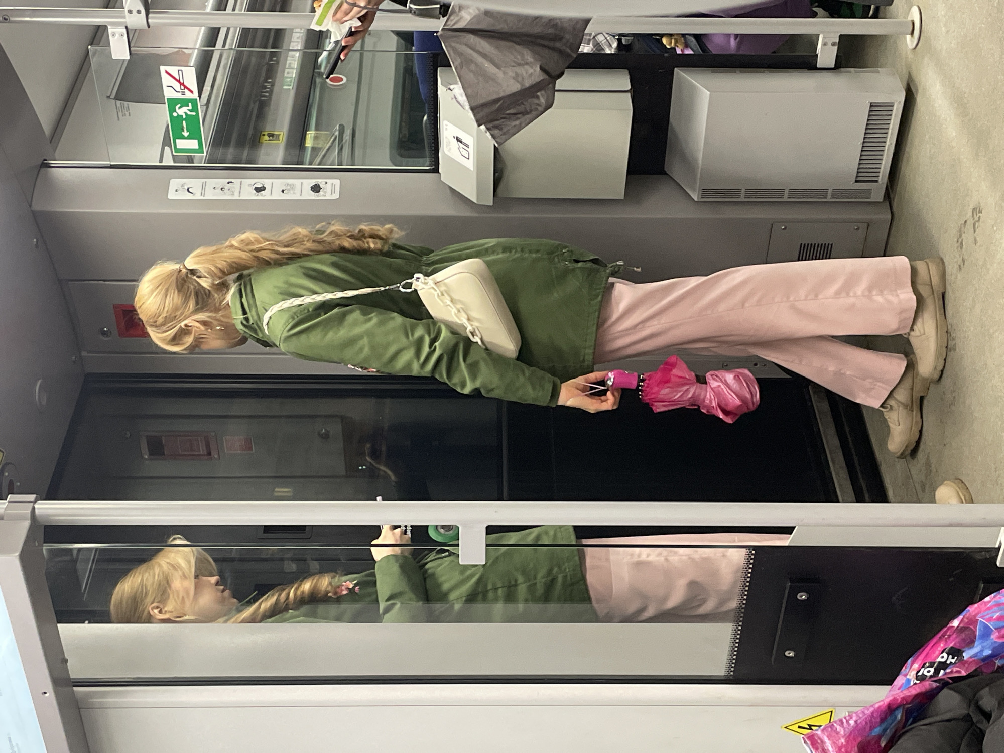 A woman with blonde hair in a ponytail is standing inside what appears to be a train or bus. She is wearing a green jacket and light pink pants. She is holding a pink umbrella. Her reflection is visible in the glass partition in front of her. Other passengers and objects are visible in the background. The location is likely within the city of Moscow, Russia, due to the design of the public transportation.