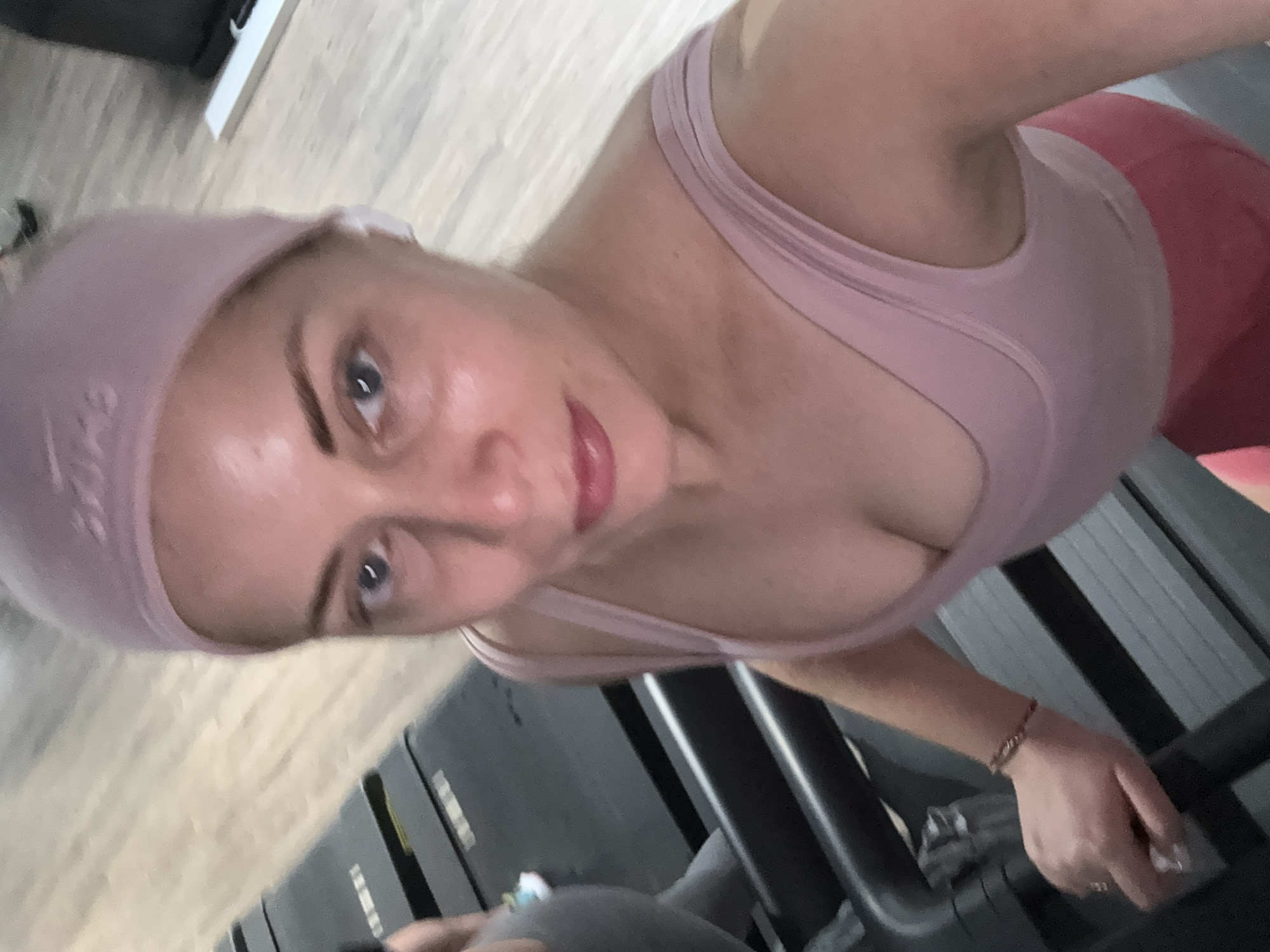 A fair-skinned woman with blue eyes and blonde hair is standing on a treadmill in a gym. She is wearing a light pink headband, a matching sports bra, and red leggings. She is holding onto the treadmill handles and appears to be taking a selfie. The gym floor appears to be wood, and there is some other equipment visible in the background. It is not possible to determine the location in Paris from this image.
