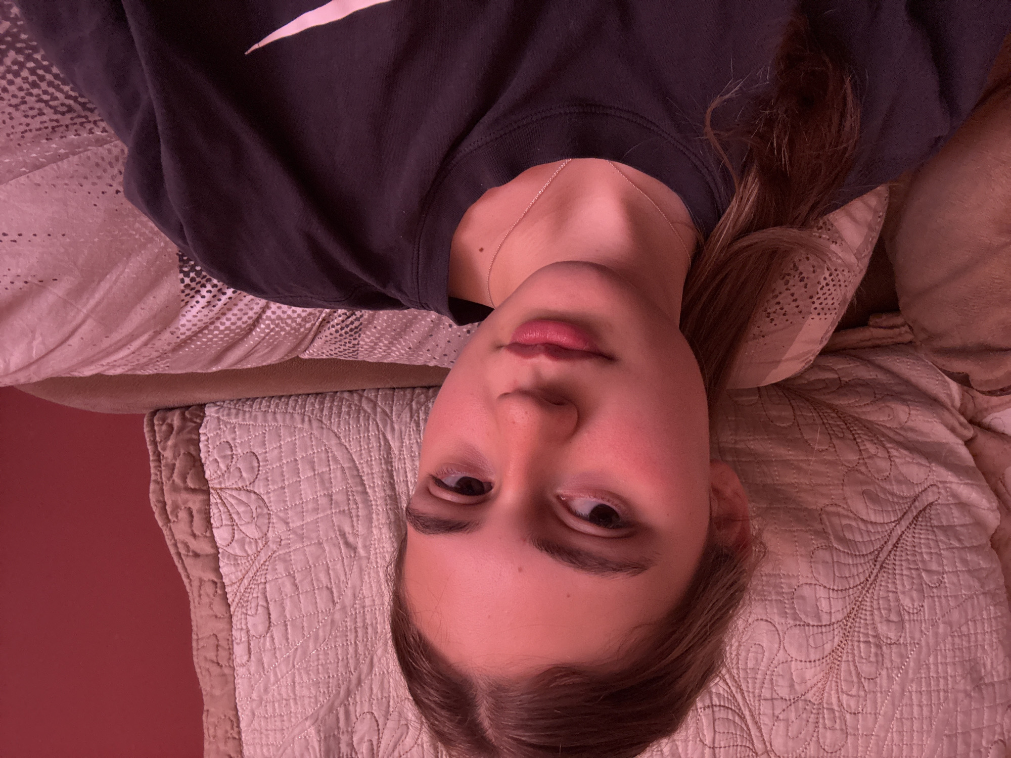 A young woman is lying on her back on a quilted bedspread and pillow. She is wearing a dark sweatshirt with a white logo visible on the upper left. She has fair skin and is wearing a thin necklace. Her hair is blonde or light brown. The background appears to be a peach or pink wall.
There is no indication from this image that the location is Panagyurishte, Bulgaria.
