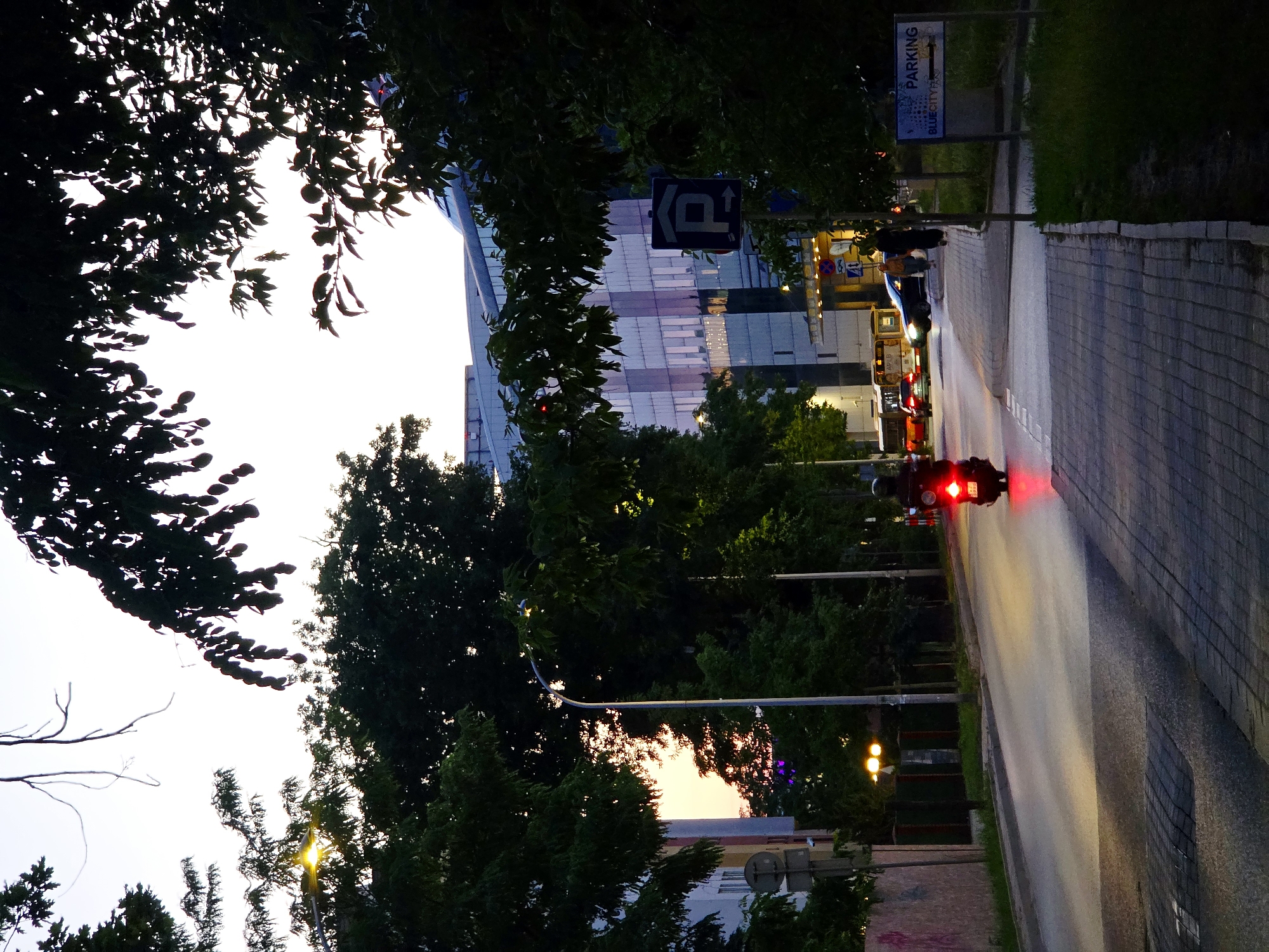 This image depicts a street scene in Warsaw, Poland, likely at dusk. In the foreground, a scooter with its red taillight illuminated is driving away from the camera on the right side of the road. The scooter is on the asphalt road, which is bordered by a cobblestone sidewalk. To the right of the sidewalk, there is a green area with grass and a sign indicating "PARKING" with an arrow pointing to the right. Behind the sidewalk and grass, trees with dark green foliage form a dense screen.
Further down the road, several vehicles are visible, including a bus and cars. A modern building with a glass facade and rows of windows stands on the right side of the street, behind the trees. The building's windows reflect the twilight sky and colors.
On the left side of the road, trees and buildings line the street. A street lamp with its light on is visible among the trees. The sky is a pale yellow and white, suggesting the last moments of daylight. The overall atmosphere is one of a city street at the end of the day, with a mix of natural elements and urban structures.