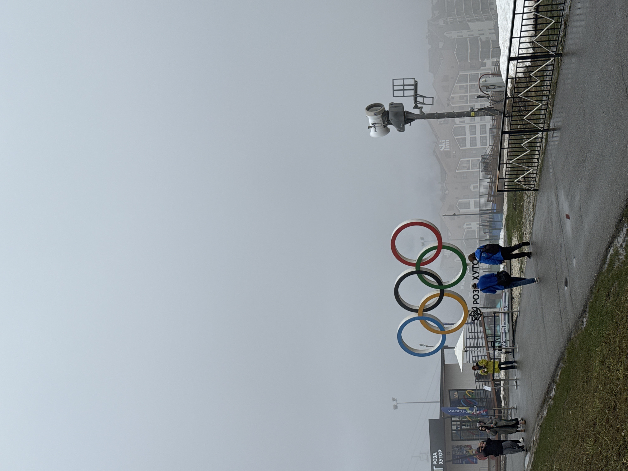 The image shows the Olympic rings in Sochi, Russia, on a foggy day. Two people are walking along a paved path next to the rings. Several people are standing in front of a building at the base of the rings. Buildings are visible in the background, obscured by fog. A black metal fence runs along the path.