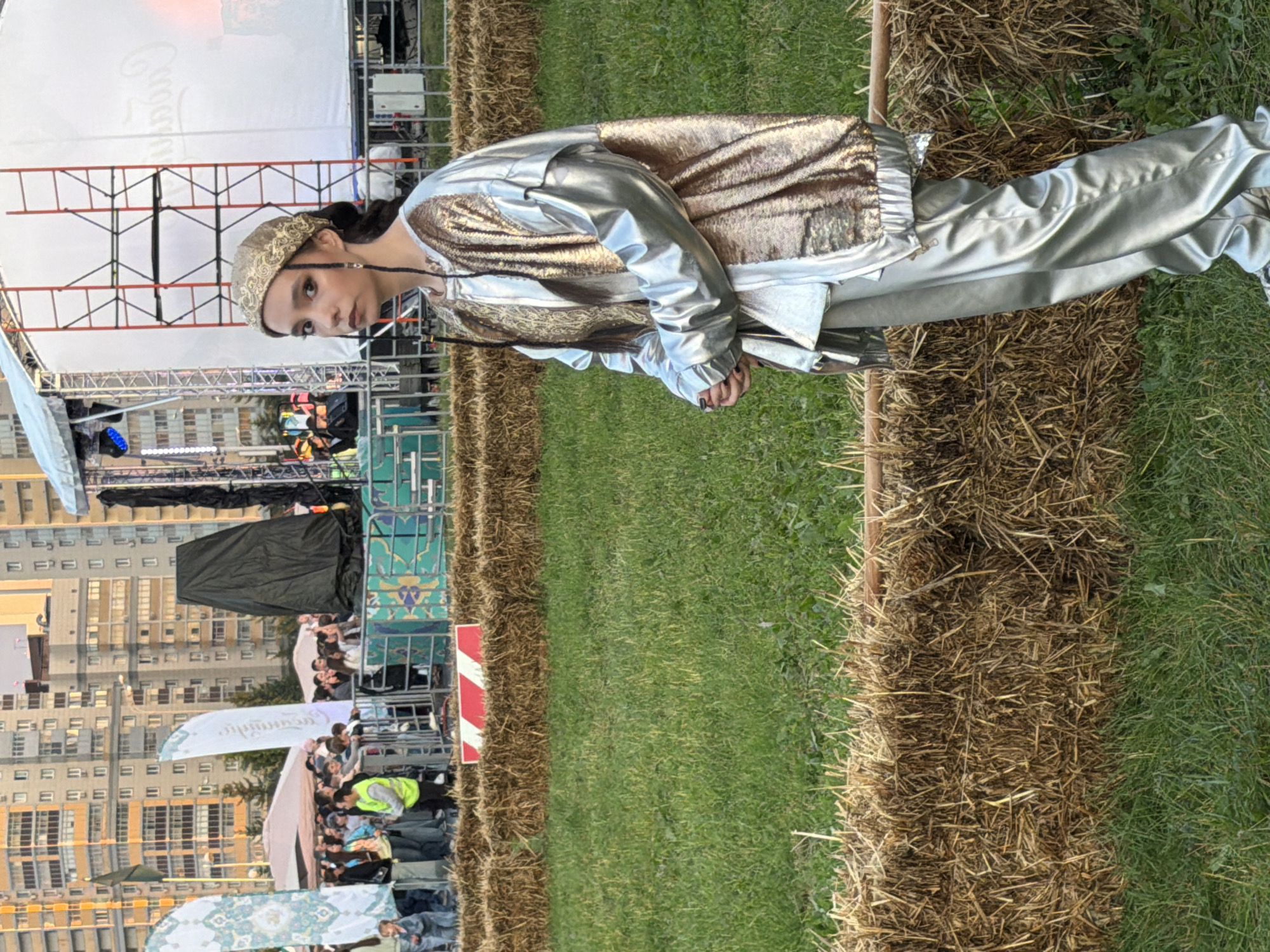In an outdoor urban setting, a young woman with dark hair styled in braids and a gold patterned headscarf stands in the foreground. She is wearing a shimmering silver and gold outfit consisting of a jacket and loose-fitting pants. Her hands are clasped in front of her. Behind her, a crowd of people gathers, some standing behind a metal barrier and others near a stage with a large white screen and scaffolding. Several flags with intricate patterns are visible among the crowd, as well as tents and tall, modern buildings in the background. The ground is covered with grass and rows of hay bales.