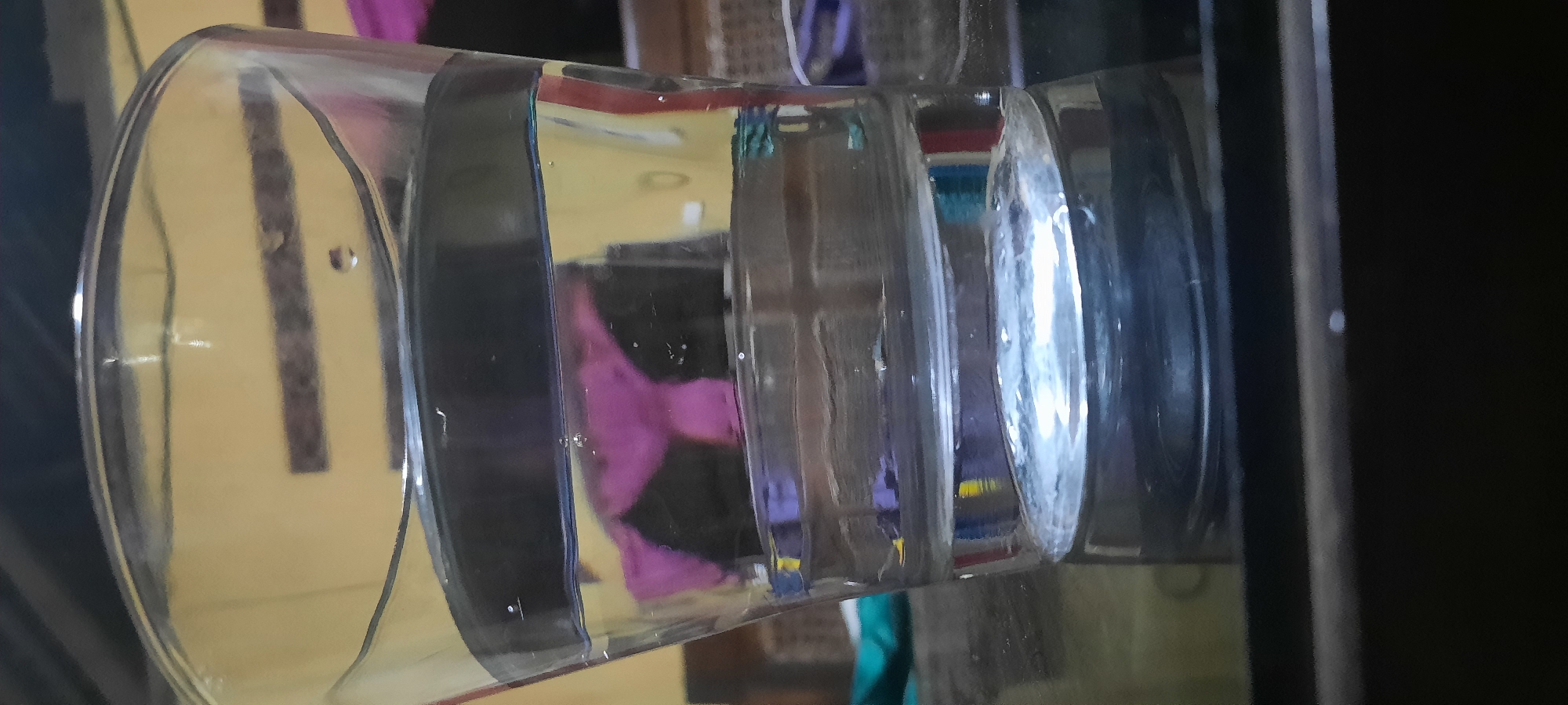 An indoor scene is depicted from a close-up, low-angle perspective, focusing on a clear glass tumbler filled with clear liquid. The tumbler is positioned on a reflective dark surface, likely a table. The liquid inside the glass exhibits a distinct stratification, with darker liquid occupying the top section and a lighter, clear liquid below. Small air bubbles are visible within the liquid.
The background of the image is out of focus but suggests an indoor environment with a yellow wall. A dark object, possibly furniture, is present in the mid-ground, and a splash of purple fabric is discernible, hinting at clothing or drapery. The lighting appears artificial, casting reflections on the glass and the surface it rests upon.
The scene is devoid of any people or overt activity. The composition is static, emphasizing the solitary glass and its contents. The overall impression is one of quietude and stillness. The location in the city of Jatitengah, Indonesia, is noted, but the visual cues within the image do not offer specific identifiers of this geographical context.