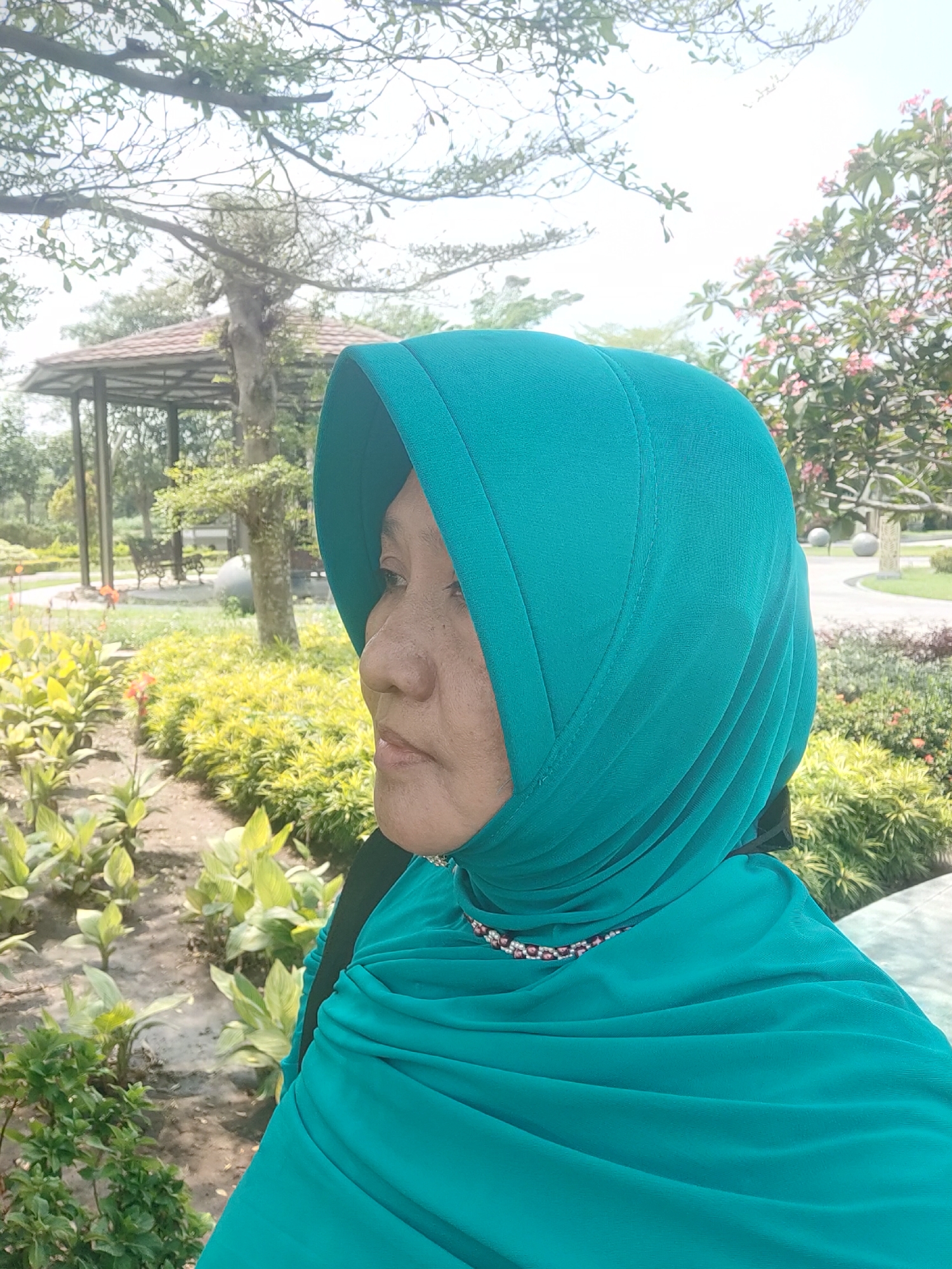 A middle-aged woman is pictured from the chest up, facing right, in a park setting. She is wearing a teal hijab, which covers her hair and neck, and a beaded necklace. Her face is partially obscured by the hijab, but her profile is visible, showing her nose and lips. Behind her, the park features lush green foliage, trees, and a gazebo with a tiled roof. The ground is covered in a dirt path and gravel. The lighting suggests it is daytime.