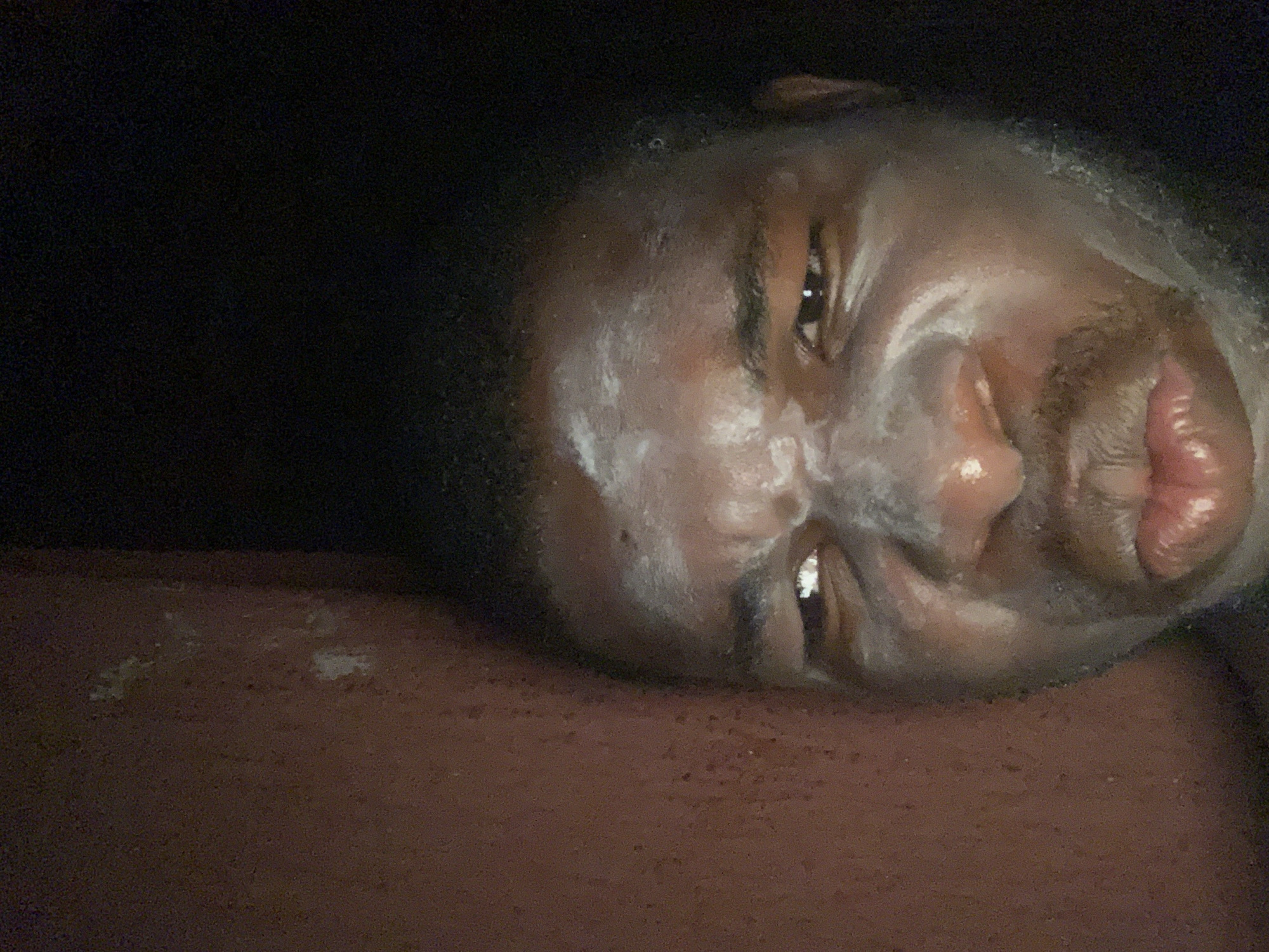 A close-up image, captured in dim lighting, features a person's face, likely situated indoors in Onitsha, Nigeria. The individual appears to be male, with short dark hair and visible stubble on the chin and upper lip. They are lying on a reddish-brown surface, possibly a bed or couch, and their face is largely covered with a thick, white, pasty substance, consistent with a facial mask or cream. The substance is applied unevenly across the forehead, cheeks, nose, and around the mouth.
The person is looking directly towards the camera with one eye clearly visible, exhibiting a subtle, perhaps knowing or amused, expression. The dim lighting creates a somewhat intimate and casual atmosphere, suggesting the image was taken at night or in a poorly lit environment. The dark background offers no specific details about the wider setting. No visible text or additional objects are present in the frame. The focus remains tightly on the subject's face and the white application.