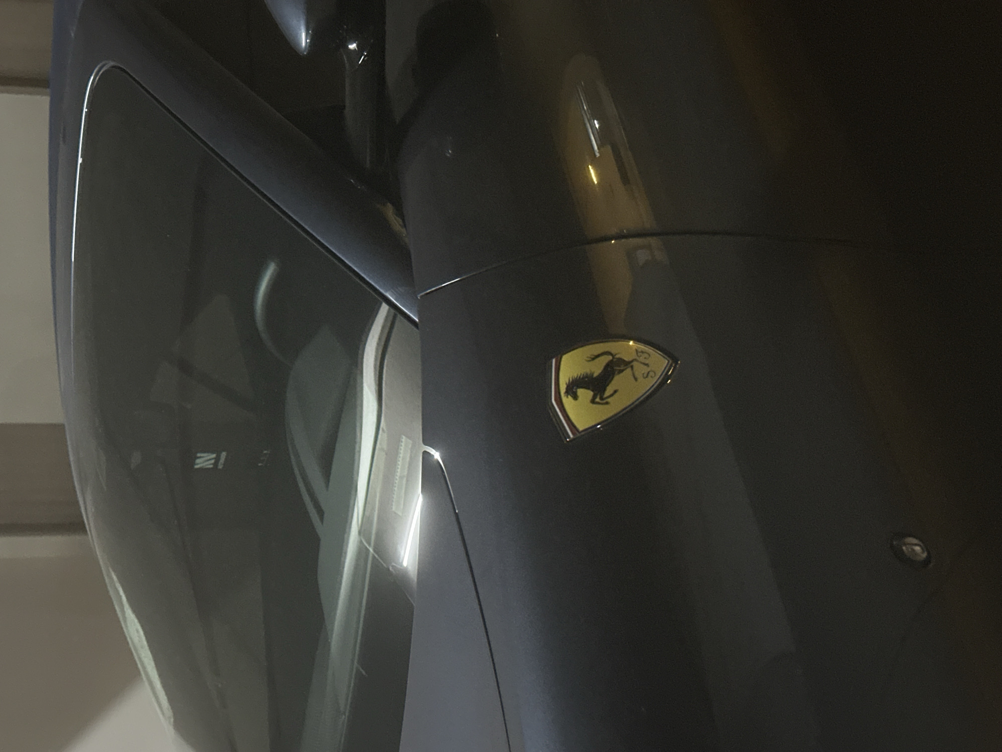The image shows a close-up of a dark-colored Ferrari car, with the iconic prancing horse logo visible on the side. Part of the windshield and the car's body are visible. The setting appears to be indoors, potentially a garage or dealership, given the controlled lighting and smooth surfaces. The location cannot be determined based on the image.