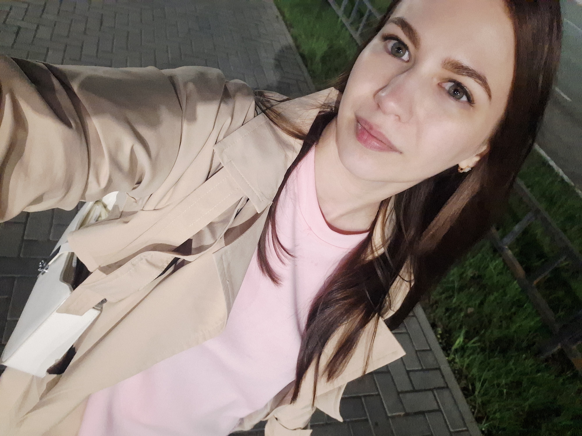 A young woman is pictured in a selfie taken on a paved walkway in Chaikovsky, Russia. The walkway is made of gray rectangular blocks. She is wearing a pink t-shirt and a beige trench coat. A white purse is also visible. Green grass and a metal railing are visible in the background. The lighting suggests it is either evening or an overcast day.