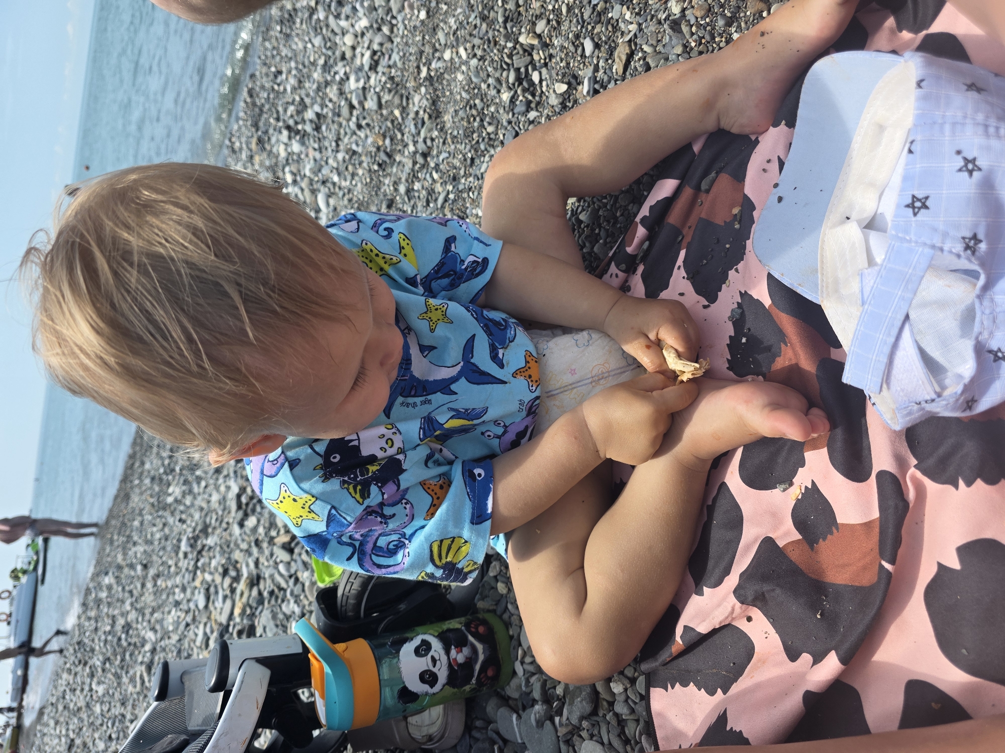 A toddler with blonde hair sits on a pebble beach, facing away from the camera and looking down at their hands. They are wearing a blue shirt with a playful sea creature print and a diaper. The toddler is sitting on a patterned blanket, possibly pink and black, with a light blue and white checkered hat resting beside them.
The scene is outdoors, with the blue sea and a slightly hazy sky visible in the background. The beach is covered in grey pebbles. A stroller with an orange and blue sippy cup featuring a panda design is partially visible on the left side of the frame. The lighting suggests it is daytime, likely sunny given the bright colors and clear water. The overall impression is of a relaxed family outing at the beach. There is no visible text in the image.