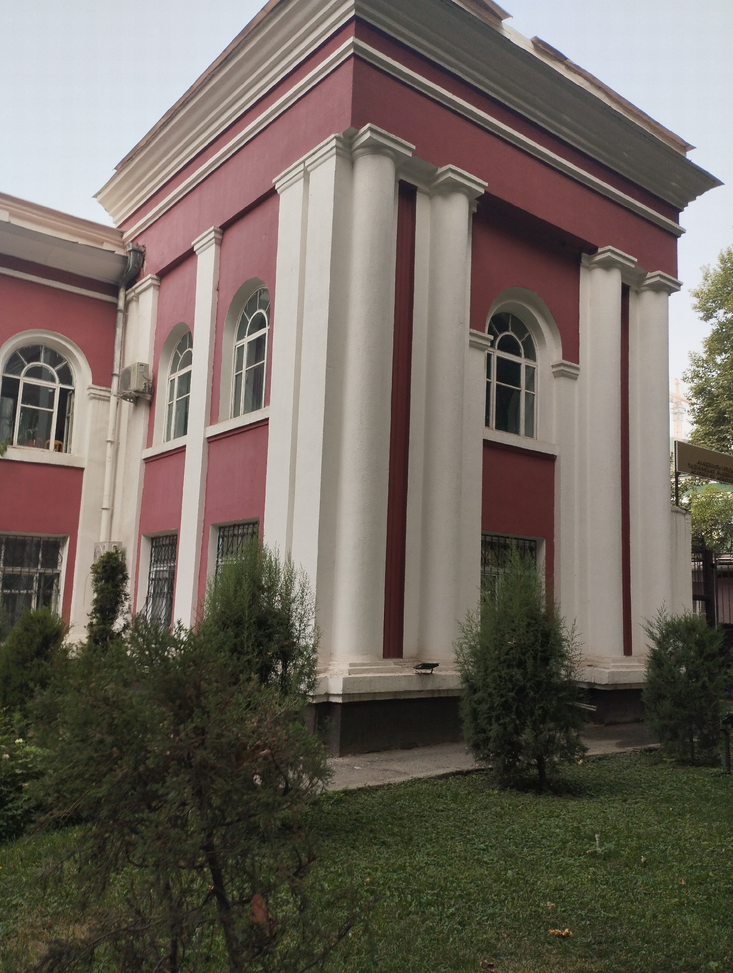 The image shows a red and white building with large, arched windows. There are white columns on the exterior of the building. In the foreground, there is a well-maintained lawn with several evergreen bushes and trees. The sky is overcast. This building is located at the National Museum of Tajikistan in Dushanbe, Tajikistan. The museum is a prominent cultural institution in the city, showcasing a variety of exhibits related to the history, culture, and art of Tajikistan.