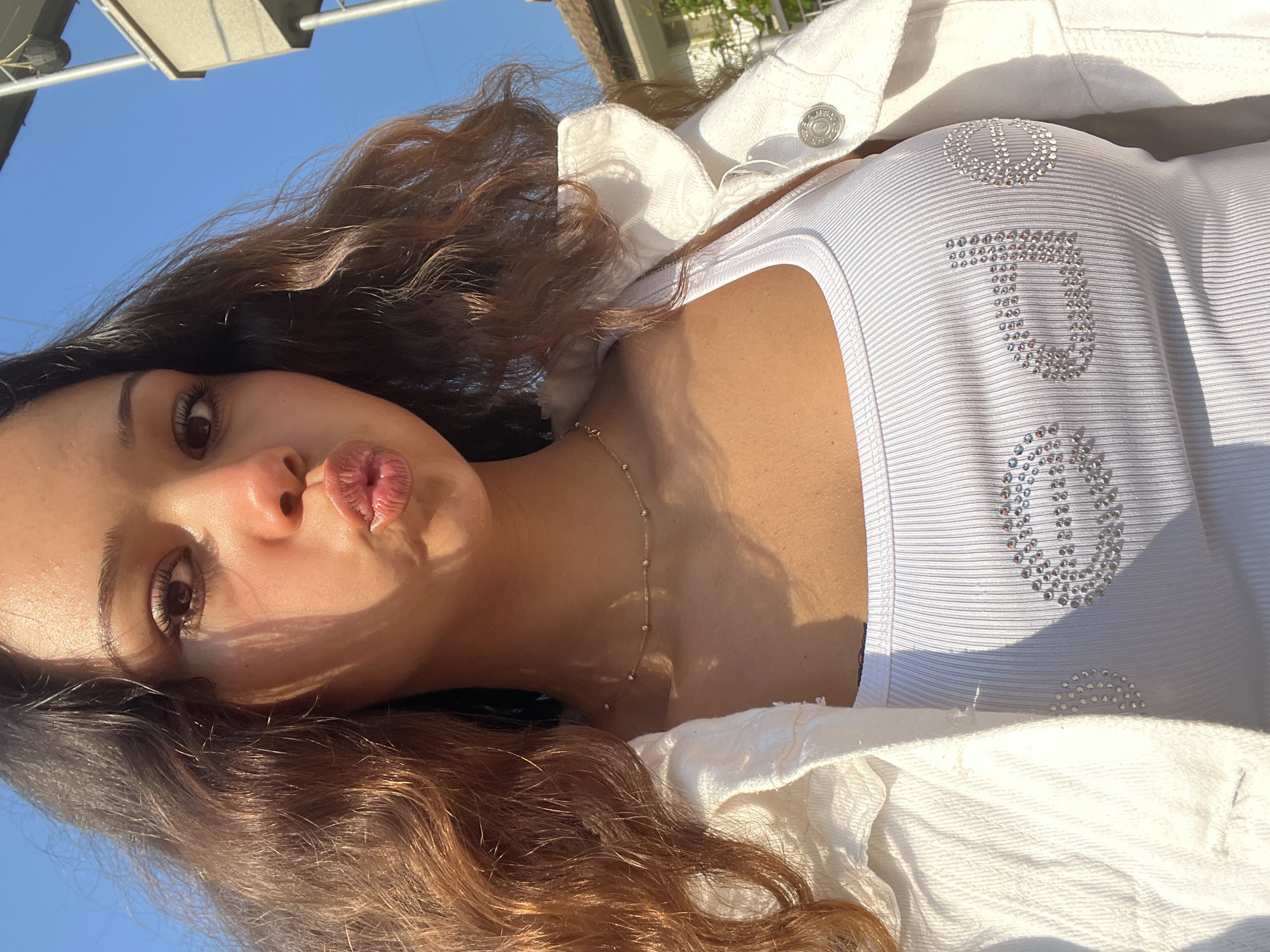 A young woman with long, wavy brown hair is depicted in a brightly lit, rotated close-up shot, likely a selfie taken outdoors. She is wearing a white ribbed tank top and a white denim-style jacket. Her lips are playfully pouted, and her right eye is visible with prominent eyelashes. A delicate gold-colored necklace with small beads is visible on her chest. The background shows a clear blue sky with some faint architectural elements or awnings. Bright sunlight casts strong highlights on her skin and clothing, creating distinct shadows.
On her white top, there are decorative patterns made of rhinestones. A peace sign symbol is clearly visible near her right shoulder. Below it, the letters "JU" are formed by rhinestones, likely part of a larger word such as "Juicy," with another partial rhinestone pattern beneath these letters. The overall scene suggests a sunny daytime setting.