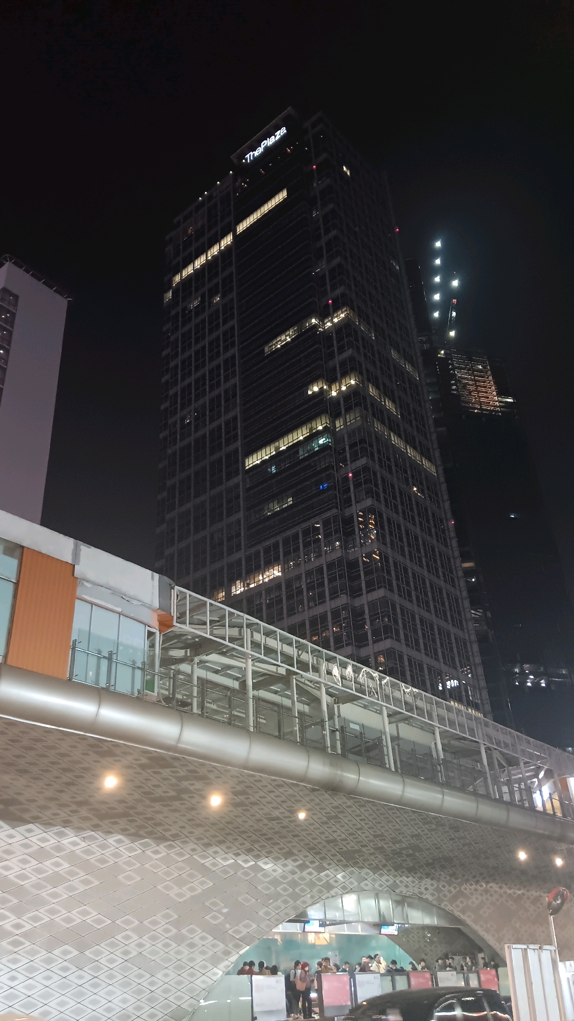 The image depicts a nighttime view of a modern cityscape in Central Jakarta, Indonesia. Dominating the frame is a tall, dark skyscraper with illuminated windows, featuring the sign "The Plaza" at its peak. In the foreground, a curved, elevated walkway or structure with a patterned underside is visible. This structure has artificial lighting illuminating its surface and the area beneath it. Below the walkway, a group of people are gathered, some appearing to be waiting or walking, with cars and traffic lights also present. Another building, lighter in color, is partially visible to the left of the main skyscraper. The overall impression is of a bustling urban environment at night, with modern architecture and public spaces.