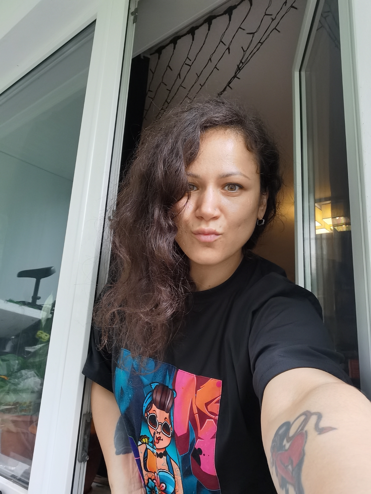 The image is a selfie of a woman looking directly at the camera with a playful, slightly pouting expression. She has long, dark, curly hair, brown eyes, a small nose piercing, and hoop earrings. She is wearing a black t-shirt with a vibrant, colorful graphic print depicting a cartoon woman in sunglasses. A tattoo with red and black elements is visible on her right upper arm.
The woman is positioned within an open window or door frame with white mullions, appearing to be taken from indoors looking out. Bright daylight illuminates her face from the left side, suggesting it is daytime. Outside the window on the left, glimpses of green plants and a dark object, possibly part of an outdoor chair or furniture, are visible. The background behind her, further inside, is darker and features decorative string lights strung across the ceiling or upper wall. The overall atmosphere is casual and direct. The location context for this media file is Chaikovsky, Russia. No discernible text is visible.