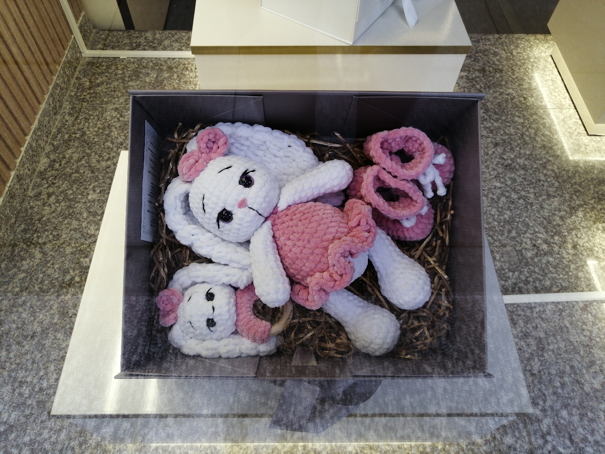 A top-down, medium shot shows two crocheted white bunny toys nestled in a dark gray box lined with shredded gold paper filler. The larger bunny is on the right, with pink crocheted ears adorned with a pink bow, a pink crocheted dress, and white crocheted feet. Its eyes are black buttons, and its mouth is a black stitch. To the left of the larger bunny is a smaller bunny toy, also white with pink accents. It has a pink bow on its head and a pink crocheted collar. Next to the bunnies, to the right, are several pink crocheted rings, likely part of a baby's toy set. The box is placed on a speckled gray and white speckled surface. The background shows a wooden paneled wall on the left and white and beige structures behind the box, suggesting an indoor setting, possibly a store or a home display. The lighting suggests an overhead light source creating some shadows. This scene is consistent with a product display in a retail environment in Batumi, Georgia.