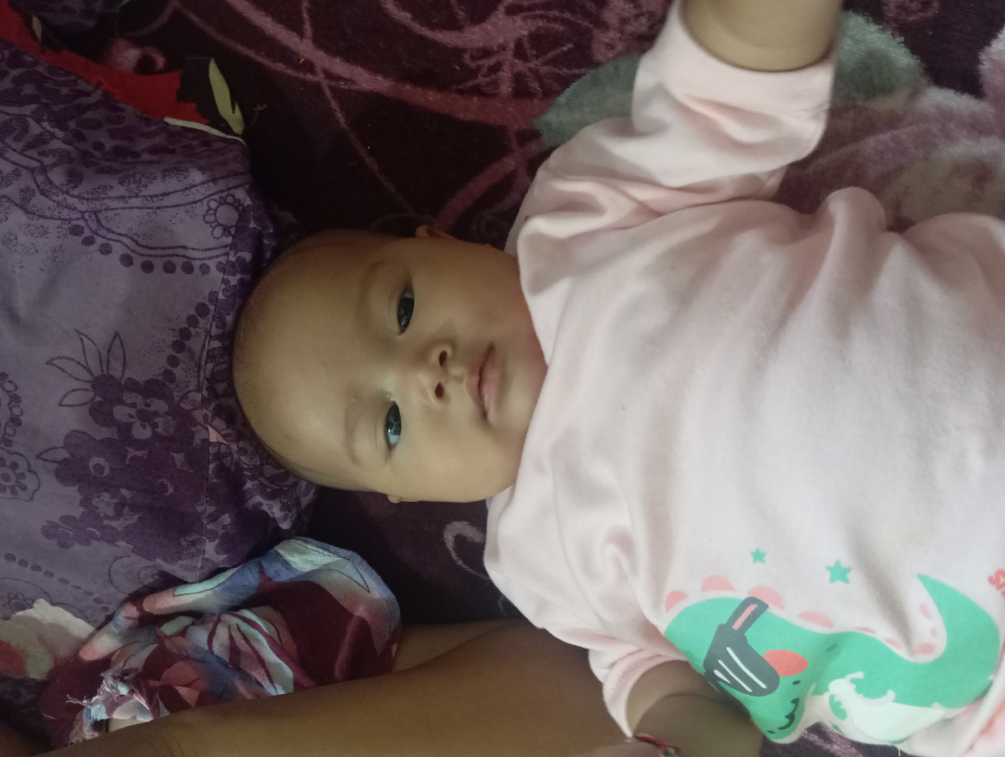 A close-up, overhead shot shows a baby lying on a patterned blanket. The baby is wearing a pale pink long-sleeved onesie with a green dinosaur graphic wearing sunglasses on the left side of the chest. The baby's head is tilted slightly to the left, and they are looking directly at the camera. The baby's left arm is visible on the left side of the frame, and their right arm is bent at the elbow, resting on their chest. The blanket underneath the baby features a dark purple floral pattern. The setting appears to be domestic, possibly a bedroom in Rejosari, Indonesia, given the context.