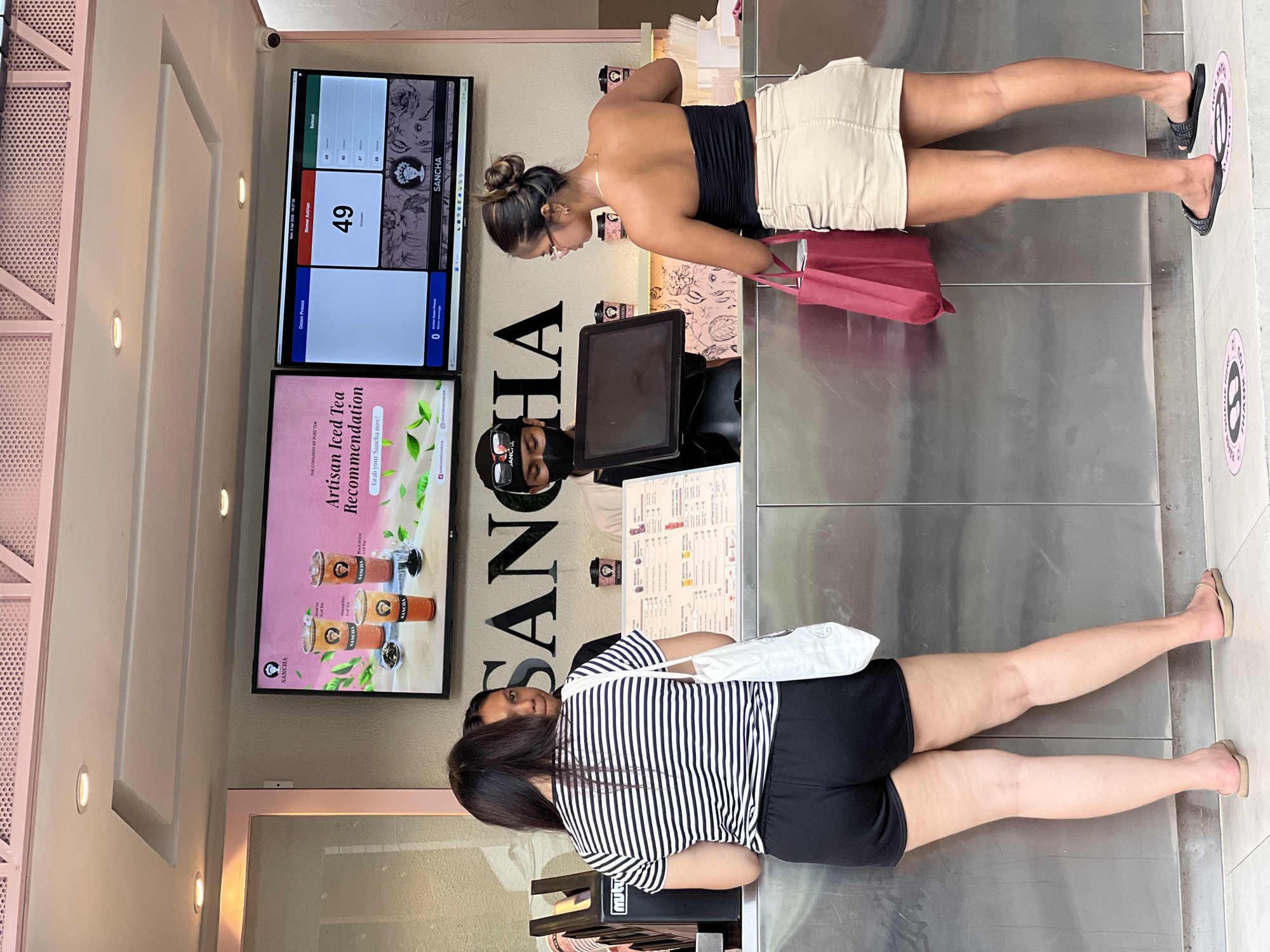 The image shows the interior of a Sancha tea shop, possibly located in Kuta, Indonesia, based on signage and the menu offering. Two female customers are standing at the counter; one in a black strapless top and light tan shorts, and the other in a striped black and white shirt and black shorts. A cashier wearing a mask is standing behind the counter. The order number displayed on the screen is 49. Advertisements for "Artisan Iced Tea" are shown on a display. Social distancing markers are on the floor.