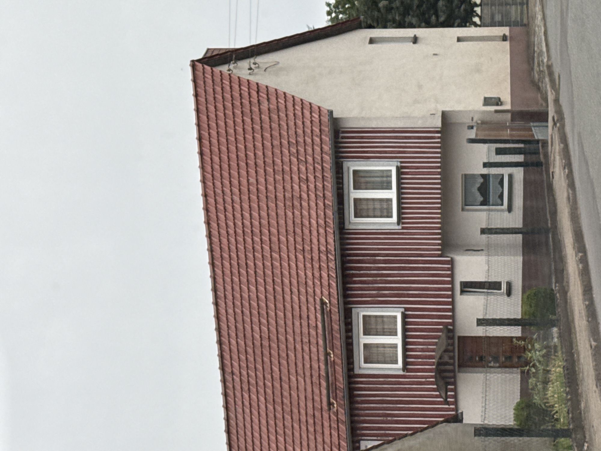 A two-story house with a red tile roof is shown from a slightly low-angle, frontal perspective in Ellrich, Germany. The house has a light-colored facade on the right and red vertical siding on the left side of the upper floor, with white trim around the windows. The upper floor has two windows on the left, both with white frames and curtains. On the right side of the upper floor, there is a single, vertically oriented window. The ground floor has a brown door with a small awning above it, and a window to its right. A fence with black posts and wire mesh runs across the front of the property, with some greenery visible behind it. The area in front of the house appears to be a sidewalk and a road. The sky is overcast.