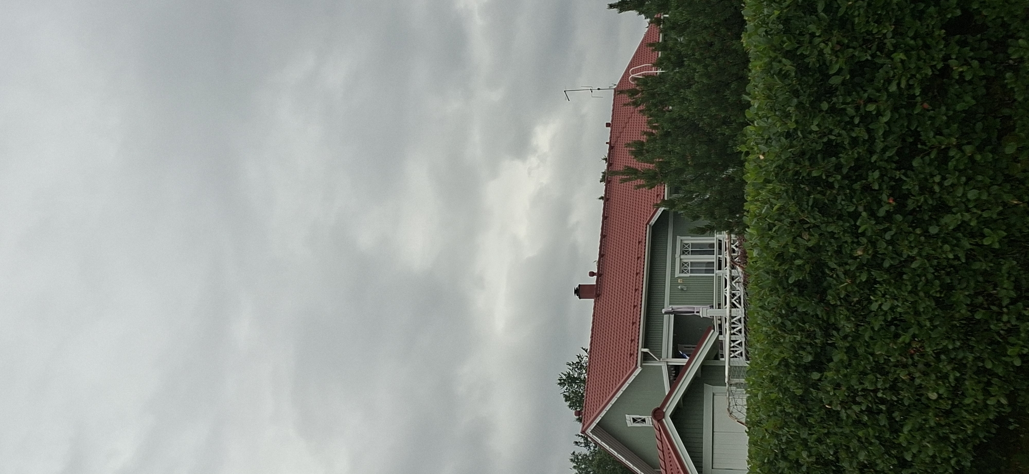 The image shows a two-story house with a red roof and green siding. The house is partially obscured by a dense green hedge in the foreground and trees in the background. The sky is overcast with gray clouds. The house has a chimney and several windows, some of which appear to be arched. There is a white railing on the second-floor balcony. The architectural style of the house suggests it is likely in a residential area of Orimattila, Finland. No people are visible in the image.