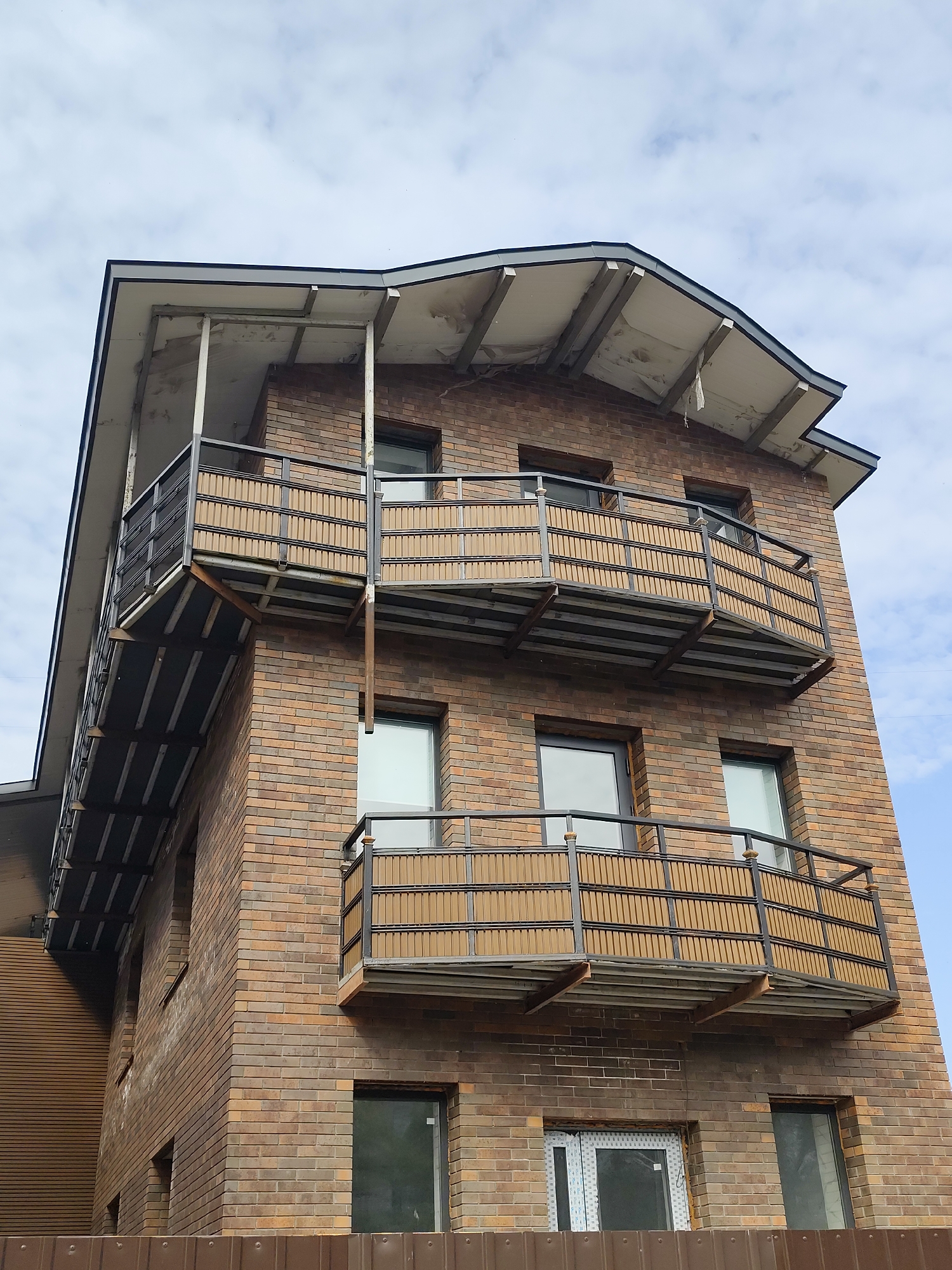 The image shows the exterior of a modern multi-story brick building under a cloudy sky. The building has a distinctive curved roofline with exposed supports. There are several balconies with metal railings and what appear to be decorative wooden slat panels. The brickwork exhibits variations in color, ranging from light brown to dark red. The windows are rectangular with dark frames. In the foreground, a brown metal fence is visible. The architecture suggests a residential or commercial structure in an urban setting, possibly part of a new development. The location is identified as Petropavl, Kazakhstan. There are no people or animals visible in the image, and no specific actions or interactions are depicted. The environment is characterized by the building and the sky.