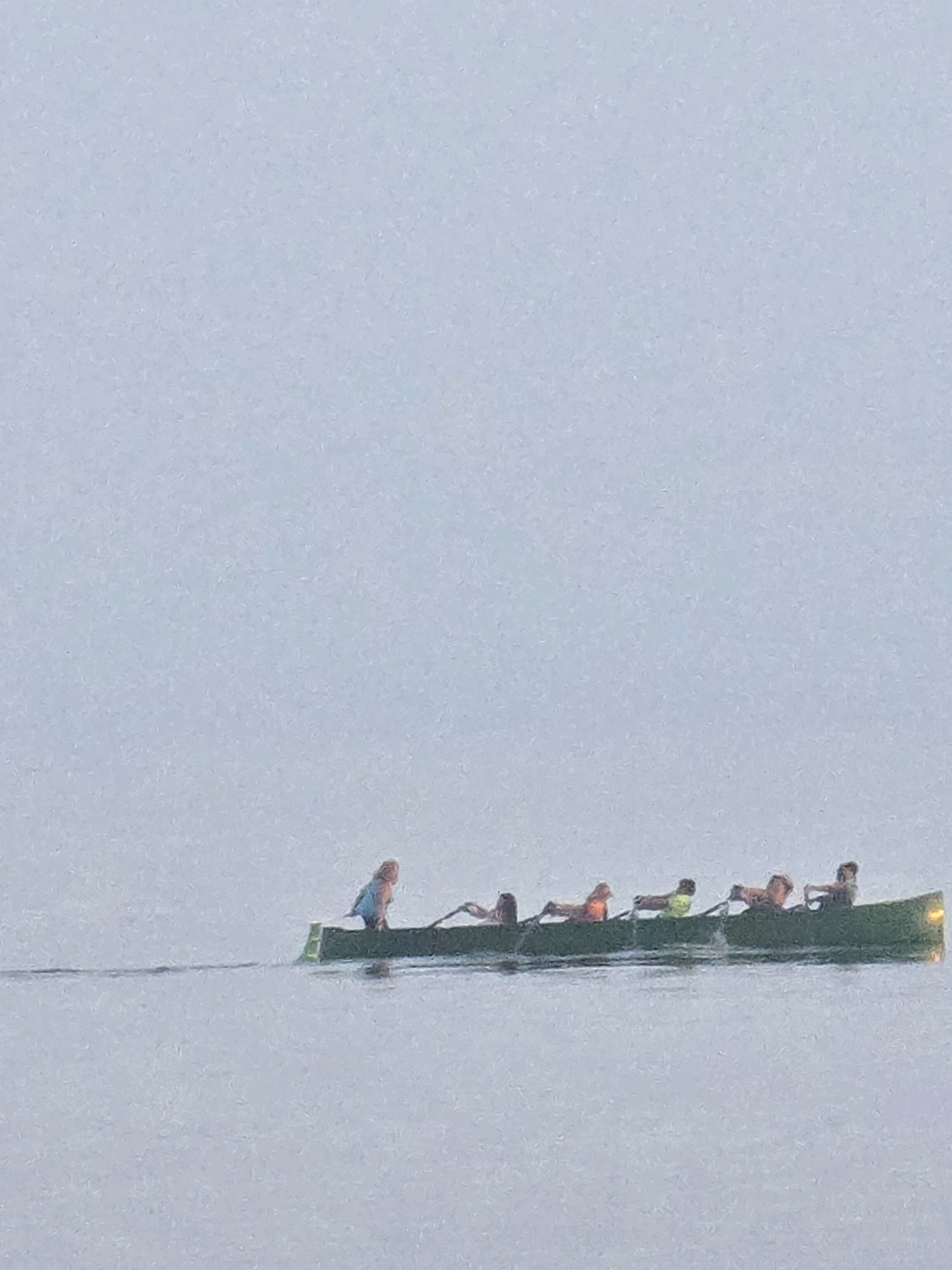 A group of six people are in a long, narrow green canoe on a calm body of water. The person at the front of the canoe, facing backward, is wearing a blue garment. The other five individuals are positioned behind, each holding a paddle. One person wears a yellow life vest. The background is a pale, hazy sky. The scene takes place on the water, potentially near the coast.