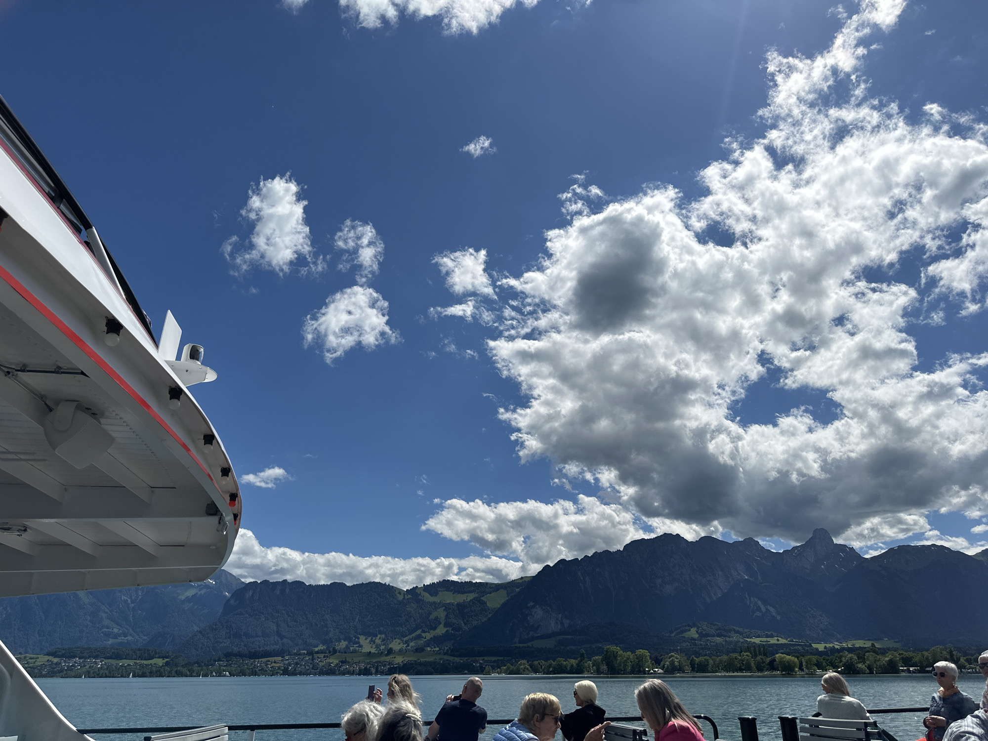 The image captures a picturesque scene from a boat on Lake Thun, Switzerland. In the foreground, the white and red structure of the boat's deck is visible, with a railing and several passengers seated on benches. The passengers, a mix of individuals, are looking out towards the lake and mountains.
The lake stretches across the middle ground, its calm, blue waters reflecting the bright sky. In the distance, a range of lush green mountains rises dramatically, their peaks dotted with snow in some areas. A small town or village can be seen nestled along the lakeshore, with buildings and trees adding to the scenic vista. The sky is a vibrant blue, filled with scattered white clouds that cast shadows on the landscape below, indicating a sunny day with some cloud cover. The overall impression is one of a serene and beautiful day in the Swiss Alps.
