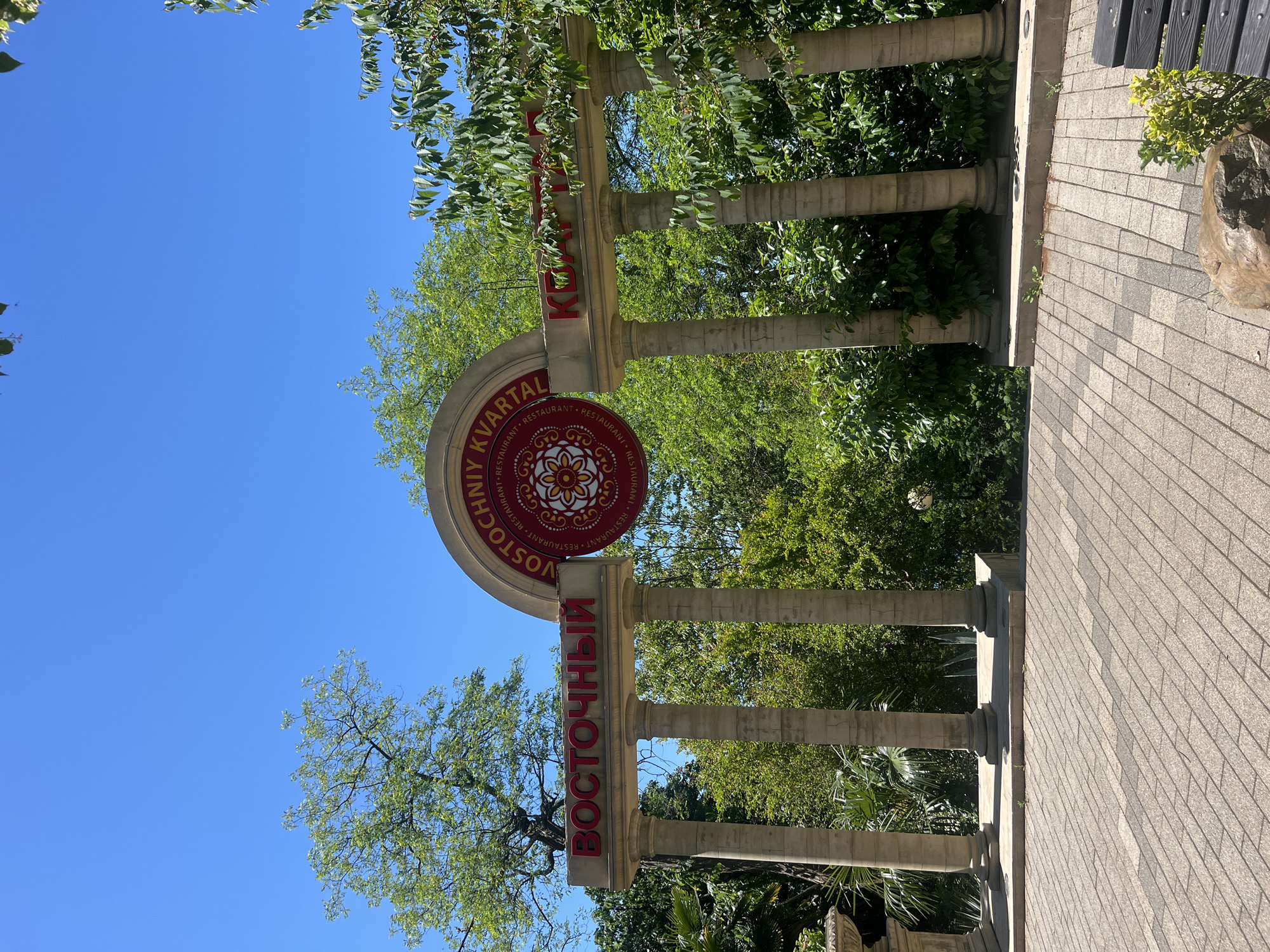 The image, oriented incorrectly and showing a clear blue sky overhead, depicts an outdoor scene under bright sunlight. It features an entrance archway or gate structure made of light-colored stone or concrete. The archway consists of a horizontal beam supported by multiple substantial columns, resembling classical architectural elements.
Suspended from the center of this archway is a large, circular sign with a red border and a central ornate pattern in red, possibly floral or geometric. The sign prominently displays text in both Cyrillic and Latin alphabets. In red letters, it reads "VOSTOCHNYY KVARTAL" (Eastern Quarter) in English transliteration, and "ВОСТОЧНЫЙ КВАРТАЛ" in Cyrillic, with smaller, repeating "RESTAURANT" text also visible in red. Further red Cyrillic text, "ВОСТОЧНЫЙ КВАРТАЛ" (Vostochnyy Kvartal), is also affixed to the horizontal beam of the archway itself.
Lush green trees and foliage are abundant, framing the archway and filling the background, suggesting a natural and possibly park-like setting. The sky is a cloudless, vibrant blue, indicating clear weather. On the right side of the image, a paved walkway or ground surface made of light grey, rectangular tiles is visible, leading towards the archway. A dark wooden bench and a large rock are partially visible on the far right, next to the tiled path. No people are present, and no specific activity is taking place. The overall impression is that of a welcoming entrance to a place, likely a restaurant or an area, given the textual cues, in Sochi, Russia.