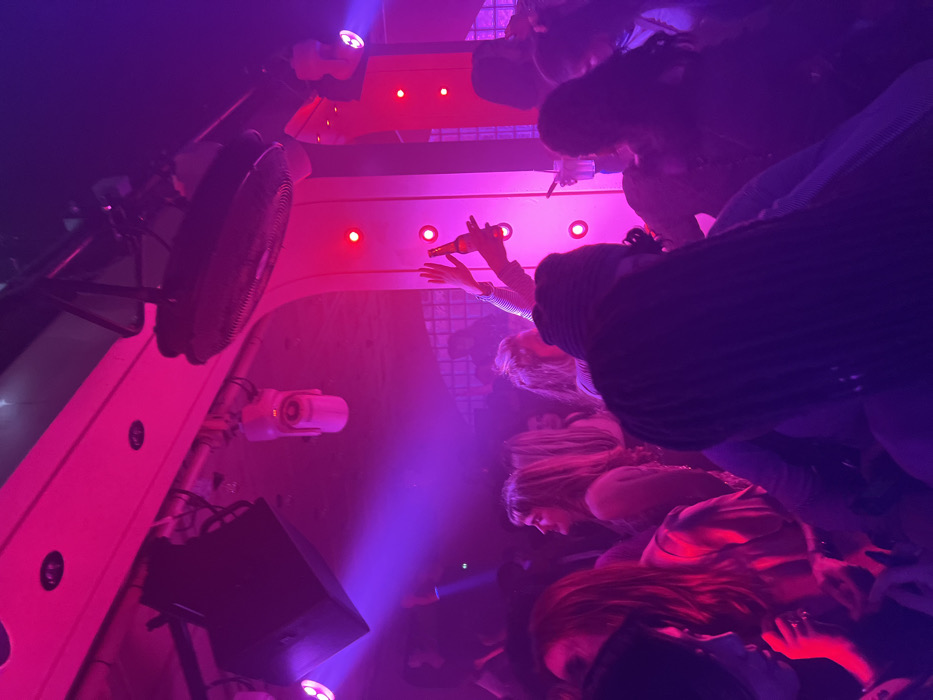 The scene depicts the interior of a nightclub or bar in Paris, France, characterized by a dim environment illuminated predominantly by magenta and purple lighting. Numerous individuals, appearing to be young adults, are present, forming a dense crowd.
In the foreground, a person with long, dark braided hair, seen from behind, faces away from the camera. To their left, another individual with long, reddish-brown hair is partially visible. Centrally, a raised arm holds a dark-colored beer bottle towards the ceiling, while another hand is also raised in the air. To the right, a person holds a clear glass with a straw.
The ceiling features exposed black speakers, various moving head spotlights emitting beams, and a large, dark industrial fan. Overhead metal trussing supports the equipment. In the background, behind the crowd, a figure resembling a DJ is visible on a slightly raised platform, positioned behind a textured or patterned glass block wall. Vertical architectural panels on the right side of the frame are illuminated with a series of circular red lights. The overall atmosphere is consistent with an active social gathering or dance event.