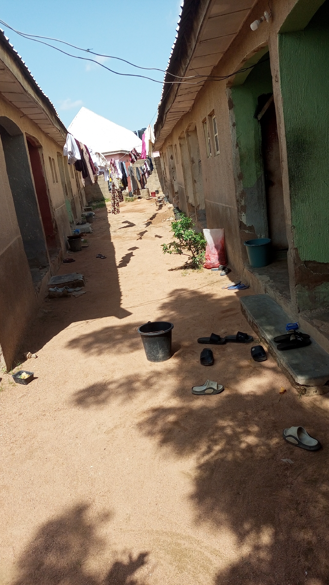 The image depicts an outdoor scene in Jalingo, Nigeria, likely a residential area or compound. Two buildings, one on the left and one on the right, line a dirt pathway. The buildings appear to be of a simple construction, with worn, textured walls. Clothes are hanging on a line strung between the buildings in the background, suggesting a laundry area.
On the ground, various objects are scattered, including several pairs of sandals and flip-flops, some black, some light-colored. A black bucket is present in the middle of the pathway, and a green bucket is visible near the entrance of the right building. A small black tray containing some yellow material is also on the ground on the left side of the pathway. A white and red plastic bag is leaning against the right building. A small green bush is growing near the middle of the pathway. The ground is covered in reddish-brown dirt or sand, with distinct shadows cast by overhead trees, indicating a sunny day. The sky is visible above, a clear blue with a few faint clouds. The overall impression is of a modest, functional living space.
