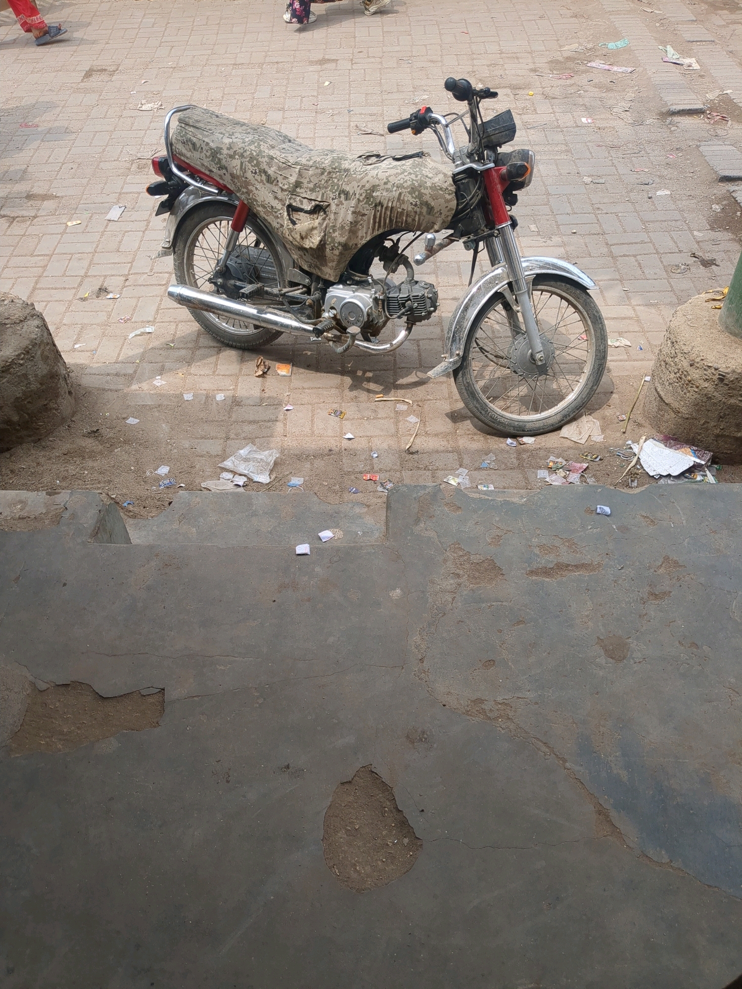 A weathered, camouflage-covered motorcycle is parked on a paved surface. The motorcycle features a chrome exhaust pipe, silver spokes on its wheels, and a red frame near the handlebars and front suspension. The seat is covered with a worn, patterned fabric, and a portion of it appears to be ripped.
The surrounding environment is somewhat littered with small pieces of paper and debris scattered on the ground. To the left of the motorcycle, a large, rough stone is visible. To the right, another large, sand-colored stone structure is present. In the background, parts of what appear to be feet of people are visible, suggesting a public area. The ground is paved with rectangular bricks, and there are areas of dirt and concrete. A concrete step or ledge is in the foreground, showing signs of wear and damage with chipped sections revealing underlying material. The lighting indicates it is daytime. The scene appears to be a street or sidewalk area in a city.
This scene is consistent with an outdoor location in Karachi Division, Pakistan, where such motorcycles are common and street environments often exhibit litter and weathered infrastructure.
