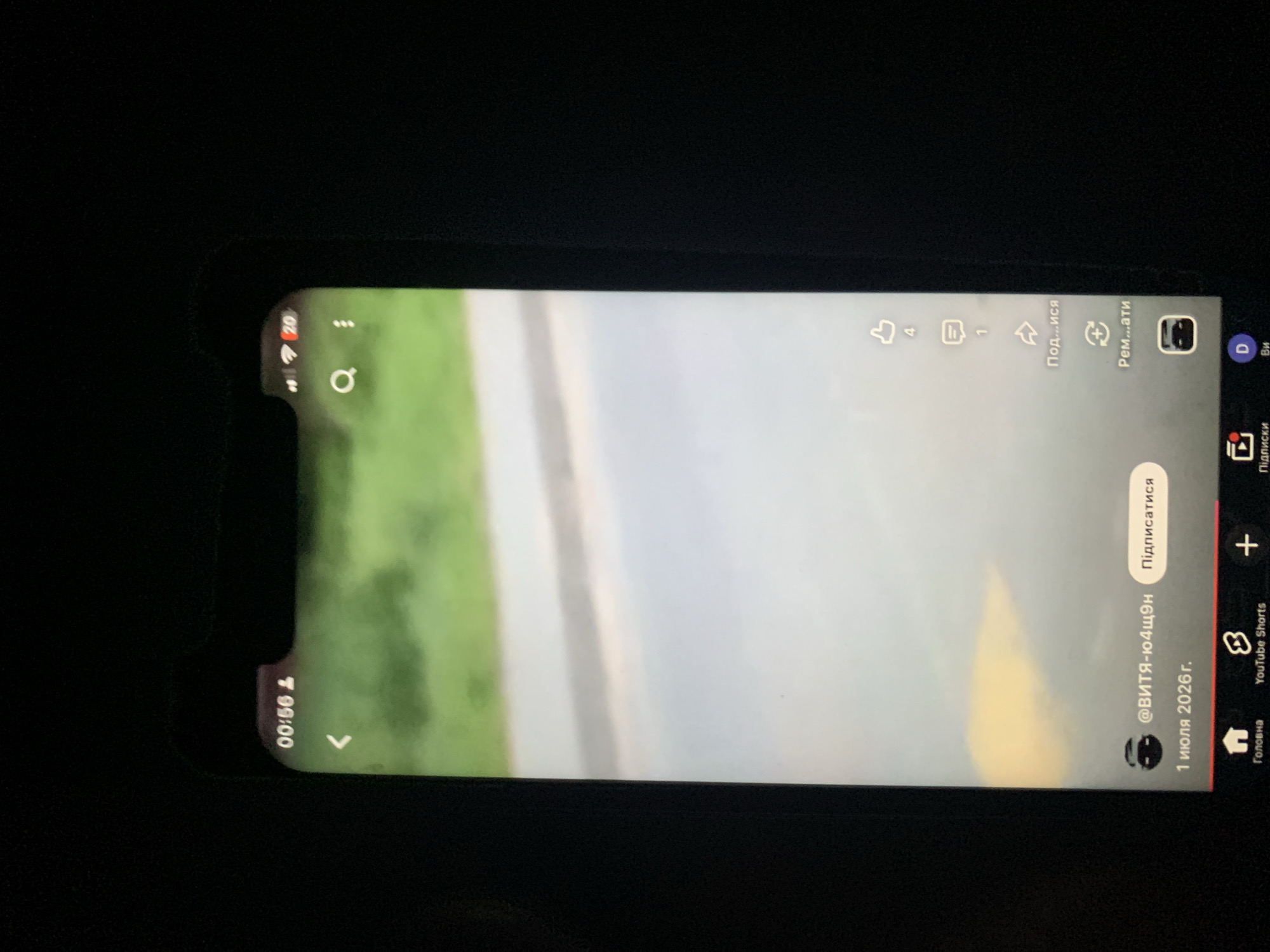 The image displays a smartphone screen displaying a YouTube Shorts video. The video features a blurred outdoor scene with greenery and a road. On the screen, there is a timestamp "00:56", a back arrow icon, a search icon, and a three-dot menu icon. Below the video, a username "@ВИТЯ-Ю4Щ9Н" is visible along with a "Підписатися" (Subscribe) button. The date "1 июля 2026 г." (July 1, 2026) is also present. On the right side of the screen, there are icons for likes (4), comments (1), sharing, and possibly a remix or reaction feature. The bottom navigation bar shows icons for "Головна" (Home), "YouTube Shorts", a plus icon for creation, "Підписки" (Subscriptions), and "Ви" (You). The smartphone is held against a dark background, suggesting it is being viewed in low light conditions. There is no information in the image that allows for a specific location in the city of Zhmerynka, Ukraine, to be identified.