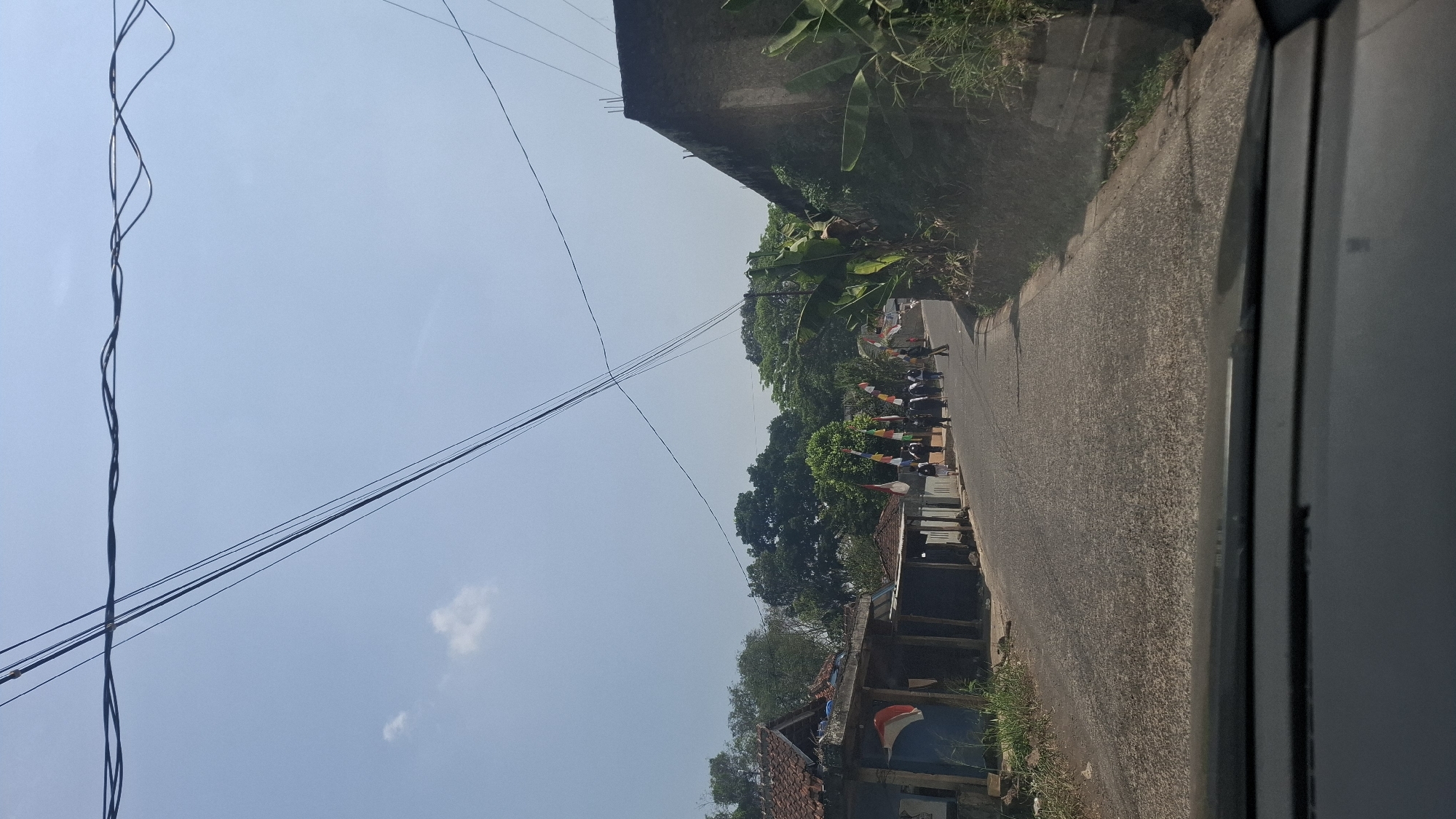 The image depicts a view from a vehicle traveling on a paved road in Cibinong Hilir, Indonesia. The sky is clear blue with a few wispy clouds. Overhead, several electrical wires are strung across the frame. On the left side of the road, a blue building with a corrugated metal roof is visible, adorned with Indonesian flags. Further down the road on the left are other simple structures. On the right side of the road, a textured concrete wall is prominent, with lush green foliage, including banana plants, growing on and around it. A group of approximately ten to twelve individuals, likely students based on their attire and demeanor, are walking in a procession along the road, heading away from the viewer. Several Indonesian flags are also displayed along the route. The road appears to be a local thoroughfare, with residential buildings and dense greenery flanking it. The overall scene suggests a community event or activity taking place on a sunny day.