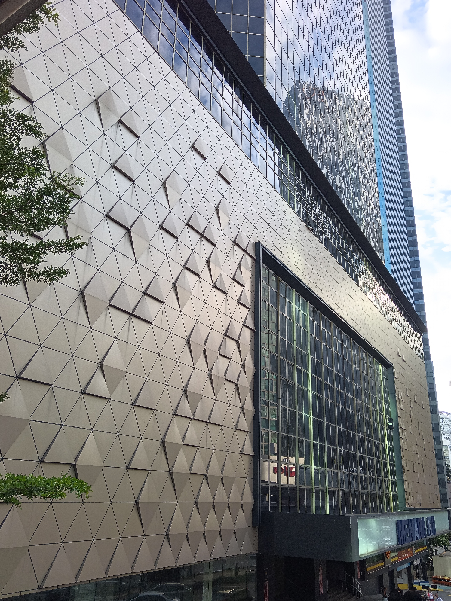 The image captures a close-up view of a modern building exterior in Kuala Lumpur, Malaysia. The building features a facade composed of triangular panels, with some panels projecting outward, creating a textured, geometric design. To the left, a portion of a tree with green leaves is visible, suggesting an urban environment with landscaping. The upper part of the building is covered in reflective glass windows, which mirror the sky and other surrounding buildings, indicating a contemporary architectural style. Below the triangular facade, a large section of the building is dominated by a grid of glass windows. At the base of the building, there are signs and store entrances, with a prominent blue sign displaying "IL BARKA" in large letters, suggesting a commercial establishment. A few people are visible near the entrance at the bottom of the frame, engaged in walking and standing. The overall scene depicts an urban street view of a commercial building in Kuala Lumpur.