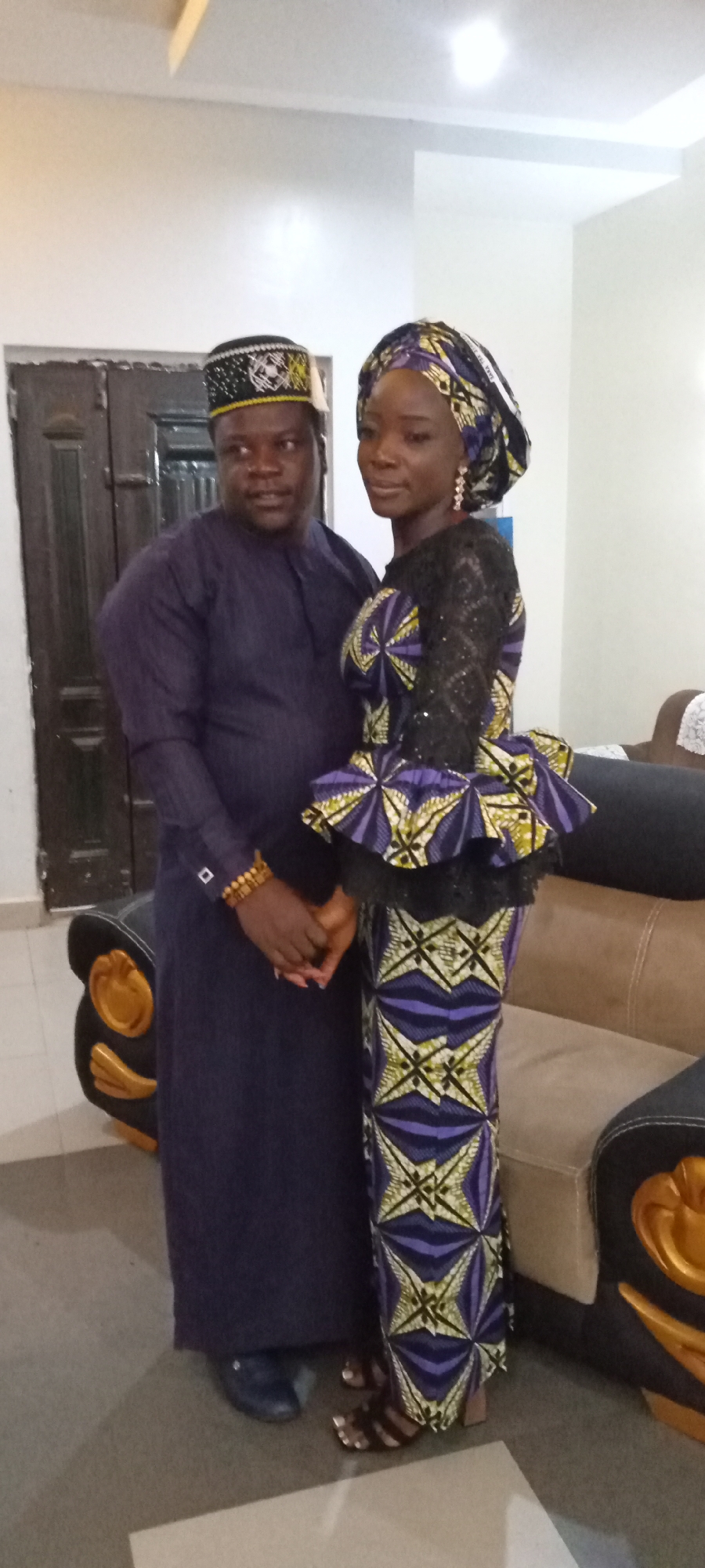A full shot shows a man and a woman standing indoors, holding hands. The man wears a dark blue, long-sleeved garment, dark shoes, and a hat with a black, white, and yellow pattern. He has dark skin and a round face. The woman stands to his right, wearing a long, patterned dress in purple, yellow, and black, with a matching head wrap. Her shoulders and upper arms are covered in a black lace material. She wears dangling earrings and has dark skin. They are standing next to a couch with tan cushions and black trim with gold accents. A dark brown door is visible behind them to the left. The location is a city of Unknown, Unknown.