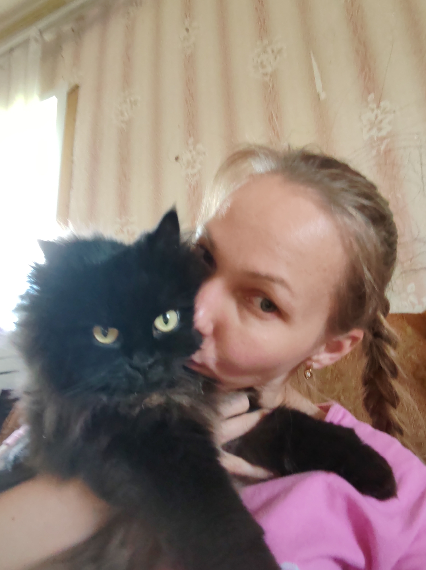 A person is holding a black cat in their arms, possibly kissing it. The person has blonde hair styled in pigtails and is wearing a pink shirt. The cat has yellow eyes. The background shows a patterned wall with a window visible on the upper left side. The location is identified as Чухлэм, Russia.