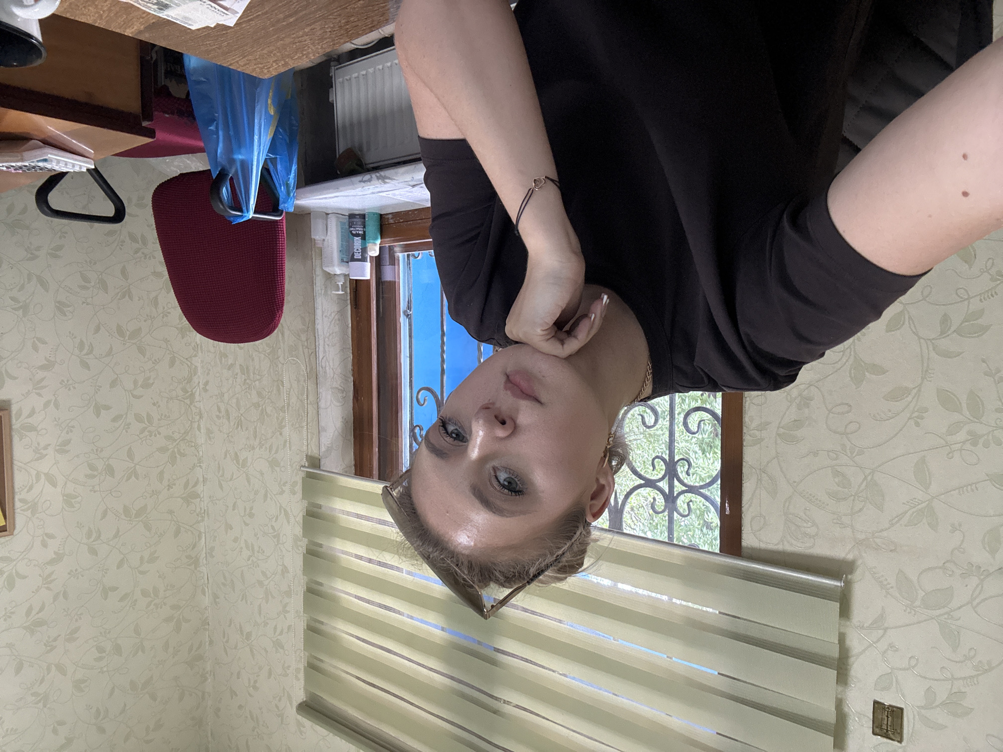 A young woman is captured in a close-up, inverted shot, her face turned towards the camera. She has light-colored skin, blue eyes, and light brown hair pulled back. She is wearing a black t-shirt and has a thin bracelet on her wrist with a heart charm. A pair of sunglasses rests on her nose. Her expression is neutral, with a slight hint of a smile.
The background suggests an indoor setting, possibly a home office or a room with a desk. To the left, a portion of a wooden desk is visible, along with a red office chair and a blue plastic bag. In the background, a window with decorative metal bars shows green foliage, suggesting it is daytime and likely pleasant weather outside. To the right of the woman, a window with horizontal blinds is visible, partially obscuring the view outside. The walls are covered in light-colored wallpaper with a subtle floral pattern. There are a few bottles of liquid and a radiator visible in the middle ground.
The image seems to be a casual selfie, taken by the subject herself. The lighting is natural, coming from the window. There is no discernible text in the image.