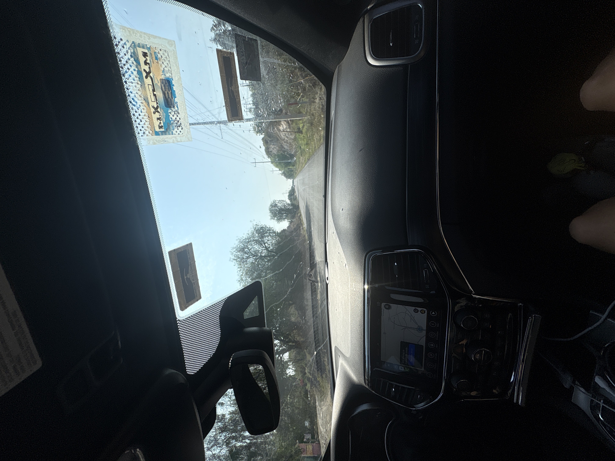 A view from inside a vehicle, possibly a car, looking out onto a road in Santiago Tlautla, Mexico. The dashboard and windshield frame the view. A navigation system is visible on the center console. Two stickers are affixed to the top of the windshield on the driver's side. The road leads into a landscape with trees and utility poles. Part of a person's legs and a green object are visible on the right side of the frame. The sky is bright with sunshine.
