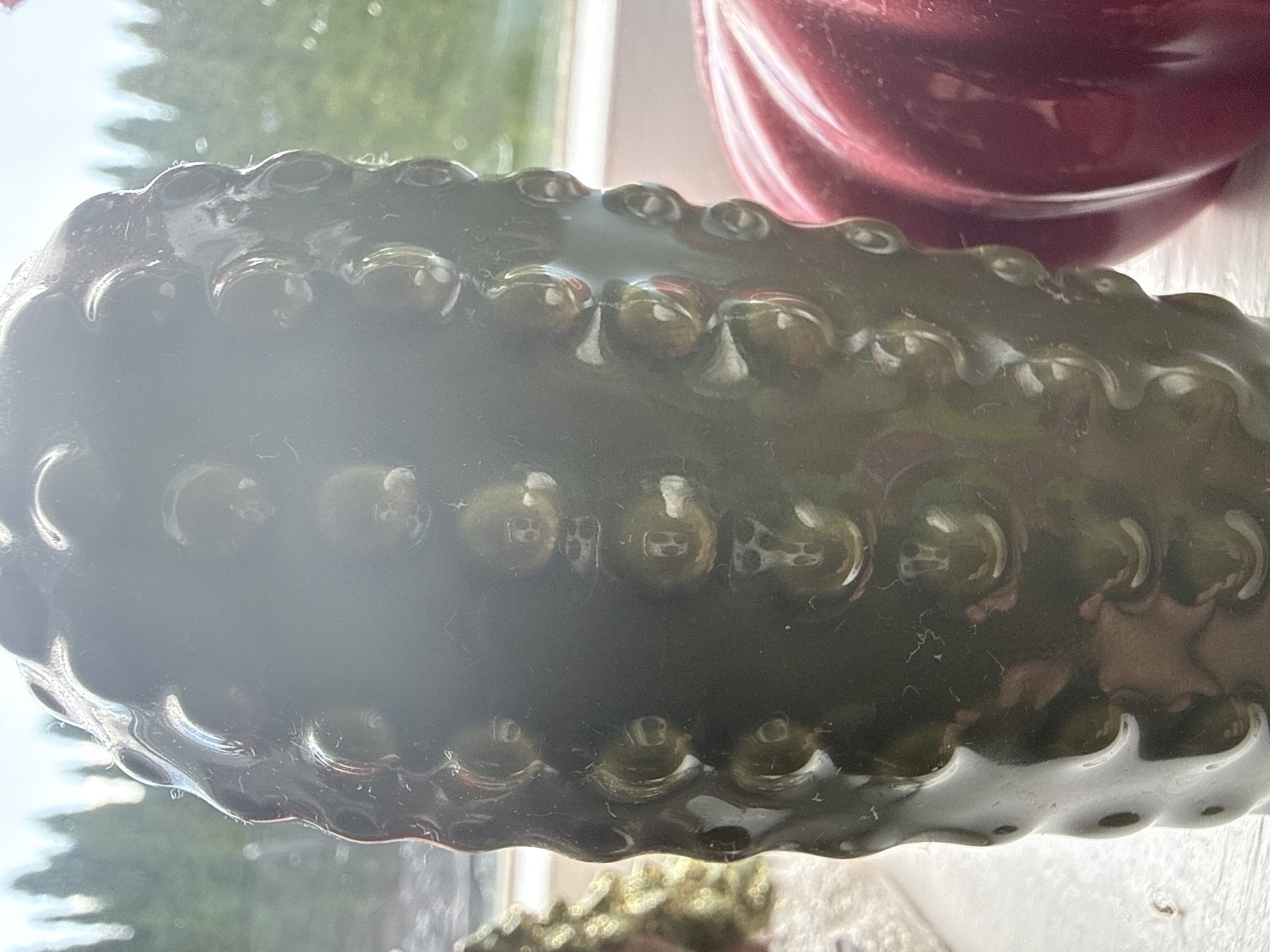 This is a close-up, indoor photograph of a dark, bumpy, candle-like object, possibly made of wax or resin, positioned in front of a window. The object is cylindrical and features a textured surface with raised, rounded protrusions. To the right of the main object, a portion of a shiny, ribbed, red object, likely another decorative item such as a pumpkin, is visible. In the background, through the window, blurred greenery and what appears to be a bright sky can be seen. The lighting suggests it is daytime. There are no people or animals present in the image. The location is stated to be in Ridabu, Norway.