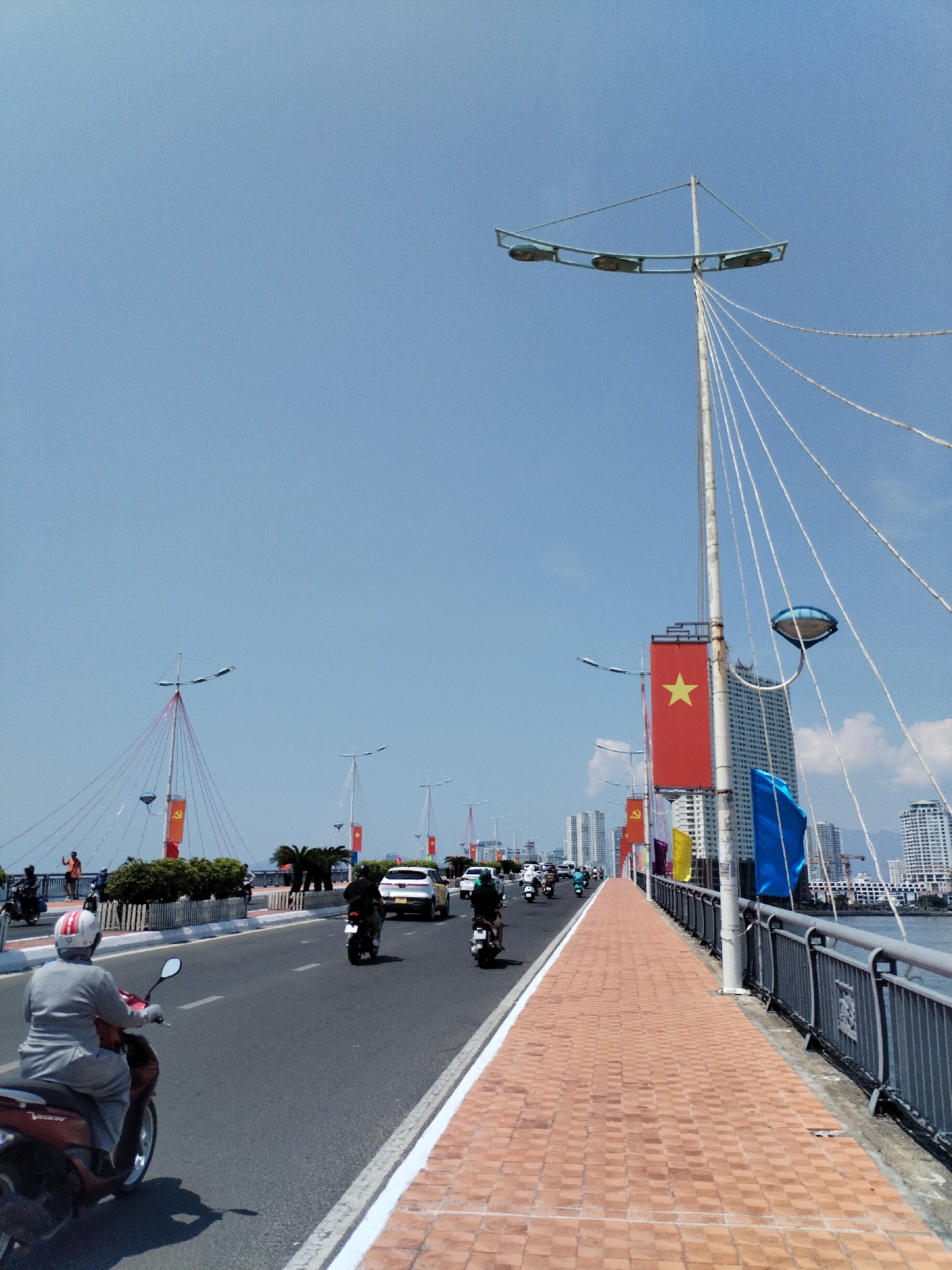 The image depicts a bridge in Bac Nha Trang Ward, Vietnam, possibly the Tran Thi Ly Bridge. The sky is clear and blue. The bridge is adorned with Vietnamese flags along its length. There is vehicular traffic, primarily consisting of motorcycles, traveling along the roadway. A pedestrian walkway runs parallel to the roadway, separated by a low barrier. Modern buildings are visible in the background, suggesting an urban setting. A person on a red motorcycle is on the left side of the image. A street light with multiple bulbs and support cables stands on the bridge.