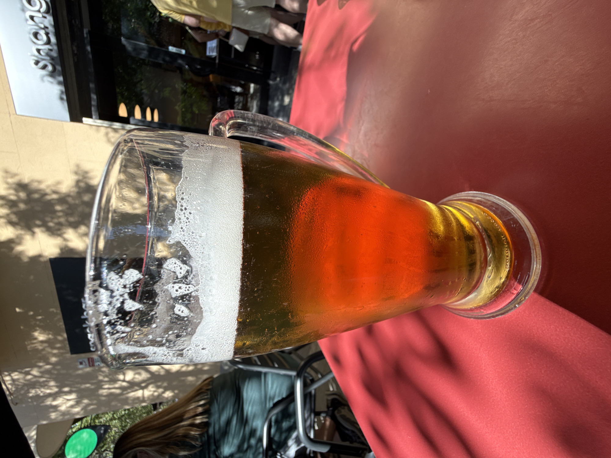 This image captures a close-up perspective of a glass of beer on a red table, likely at an outdoor cafe or restaurant terrace in Barcelona, Spain.
In the foreground, a large, clear glass pint of amber-colored beer is positioned centrally on a vibrant red tablecloth or table surface. The beer has a substantial white head of foam, and condensation droplets are visible on the exterior of the glass, indicating a cold beverage on a warm day. Distinct, dark shadows of leaves or an overhead structure are cast across the red table, particularly on the left and right sides.
To the left of the beer glass, a person with long, light brown hair is visible from the back, wearing a dark green top, seated at another table with a black metal chair partially in view. Behind them, on a light beige building wall, an indistinct dark sign with white text is partially visible, reading "snack bar". A small red and white sticker, possibly a security alarm sign, is attached to the wall below the "snack bar" sign, with the word "ALARM" discernable. Further to the upper left, a bright green circular object, possibly a light fixture or sign, stands out against the building facade. The beige building wall also displays clear shadows of foliage from an overhead tree.
To the right of the beer glass, a modern building facade with dark, reflective glass is visible. Prominently displayed on this facade, in silver-colored lettering, is the word "Shang", likely part of a restaurant or business name. Reflections on the dark glass facade show hints of trees and sunlight. Three horizontal, warm-toned lights are reflected in the glass, suggesting interior lighting or decorative elements. Partially visible near the entrance of this modern building are the lower body and legs of a person wearing a yellow t-shirt and light-colored shorts, along with another person's legs, suggesting people walking or standing nearby. One person on the right is holding a white handbag or object. The overall scene depicts a casual outdoor setting on a sunny day in an urban environment.