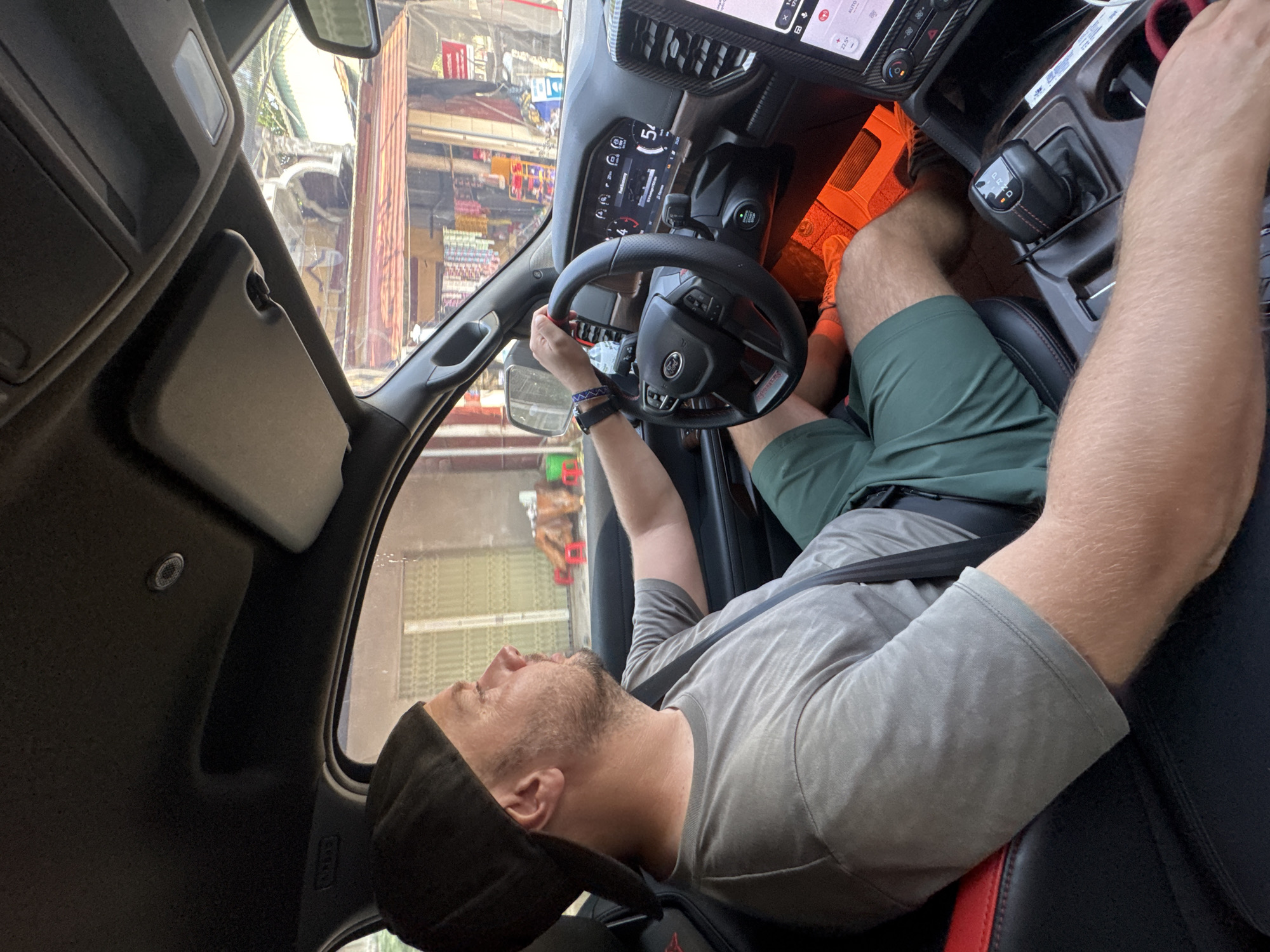This rotated image captures an interior view of a vehicle, looking towards the driver and the road ahead. A man is seated in the driver's seat, wearing a gray t-shirt, green shorts, and a black baseball cap. He has a beard and is buckled in with a seatbelt. His right hand rests on the black and red accented steering wheel, which features a "RAM" logo. His left arm is extended upwards towards the car's roof.
The dashboard's digital display shows "54", the time "11:51", and temperature readings "17.0°C" and "23.5°C". The center console includes a gear shifter clearly indicating "P R N D". Orange ambient lighting illuminates the footwell area.
Through the windshield, a bustling street scene unfolds under bright daylight. Buildings, storefronts, and what appear to be market stalls with various goods are visible, indicative of an urban or semi-urban environment. These visual cues are consistent with the location of Thông Nguyên Commune, Vietnam. The man's gaze is directed upwards, suggesting observation or rest. No specific emotion is clearly discernible. The overall impression is a moment captured inside a vehicle in a lively street setting.
