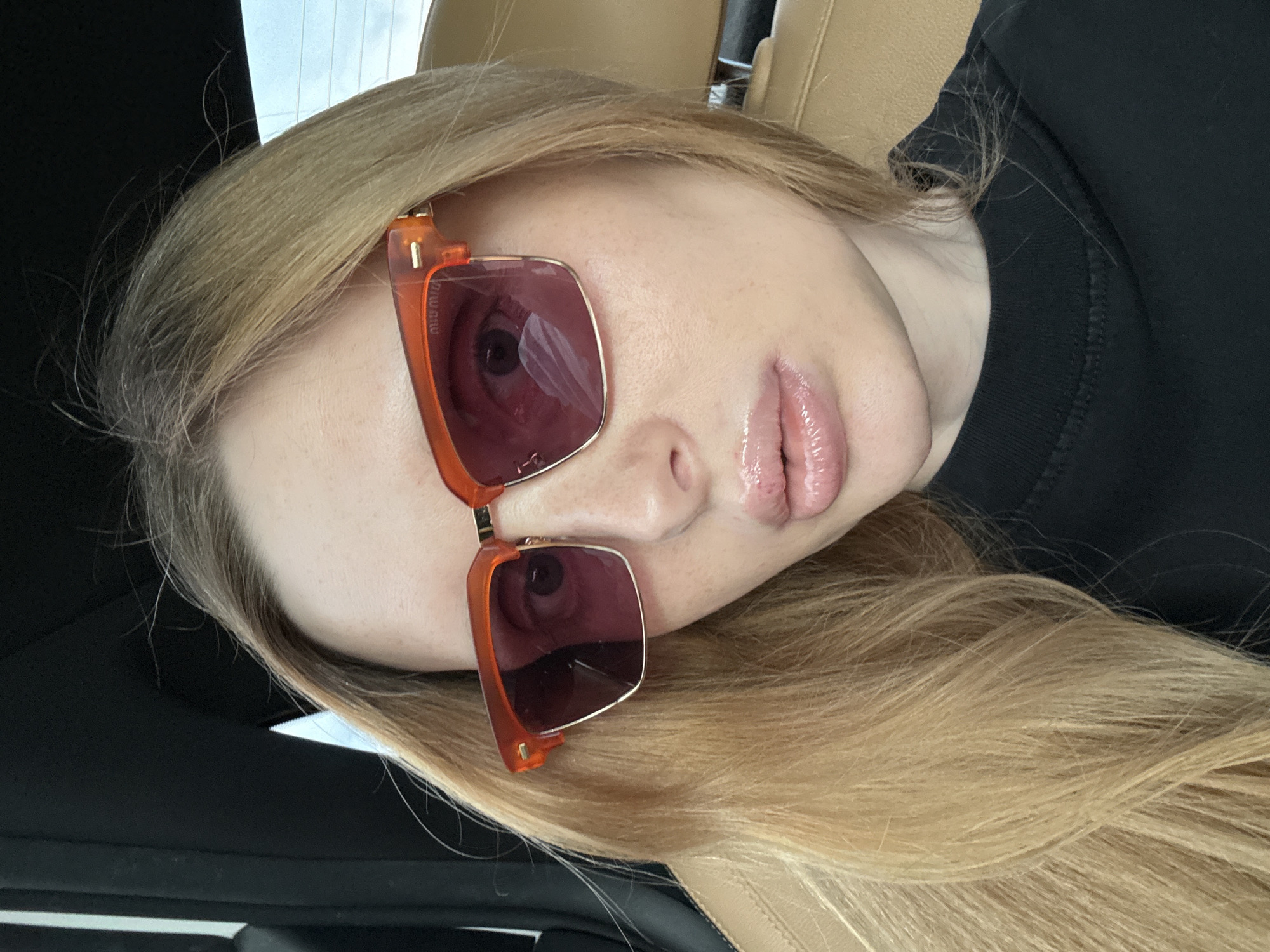 A close-up, waist-up, indoor selfie shows a Caucasian woman with blonde hair and fair skin, wearing large orange-rimmed sunglasses with pink lenses. She is wearing a black t-shirt and is seated in a car, as indicated by the car seat visible in the background. The lighting suggests it is daytime. The woman's lips are slightly parted and appear to have lip gloss.