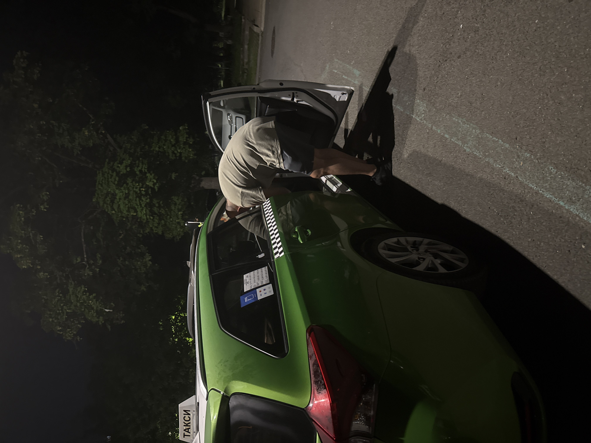The image depicts an outdoor nighttime scene in Burgas, Bulgaria, centered around a green taxi car and a man. The man, viewed from behind, wears a light-colored t-shirt and dark shorts. He is bent over, deeply leaning into the open front passenger door of the green vehicle, seemingly searching for something or interacting with the car's interior.
The car is a bright green taxi, distinguished by a white and black checkerboard decal running along its side. A prominent white sign on the roof clearly displays "ТАКСИ" in black Cyrillic letters. On the rear passenger window, several stickers are visible; one blue sticker, possibly for a payment service, shows "BGO" and other details, while additional text and symbols likely provide information about the taxi service.
The vehicle is parked on an asphalt surface, where a faint light blue chalk or paint line can be seen. In the background, dark foliage, likely trees or bushes, is visible against the night sky. The scene is illuminated by artificial light sources, casting shadows and highlighting the immediate area around the car. The overall darkness confirms that the event is taking place at night.
