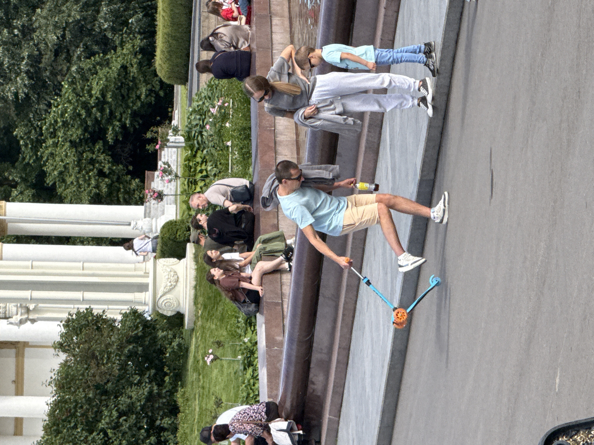 The image depicts a sunny outdoor scene in Moscow, Russia, likely a park or public square. In the foreground, a man in a light blue t-shirt and khaki shorts is bent over, holding a bottle of beverage and operating a blue scooter with orange wheels. To his right, a woman in a gray t-shirt and white sweatpants stands with her hand on her hip, holding a gray jacket. Beside her, a young boy in a light blue t-shirt and blue jeans stands looking towards the scooter.
In the mid-ground, several people are seated on a stone ledge. Some are facing away from the camera, while others are looking towards the foreground. A fountain or water feature is visible behind the seated individuals.
The background features a white classical-style structure with columns and arches, partially obscured by lush green trees and bushes. The overall atmosphere suggests a relaxed, leisurely day in a well-maintained public space.