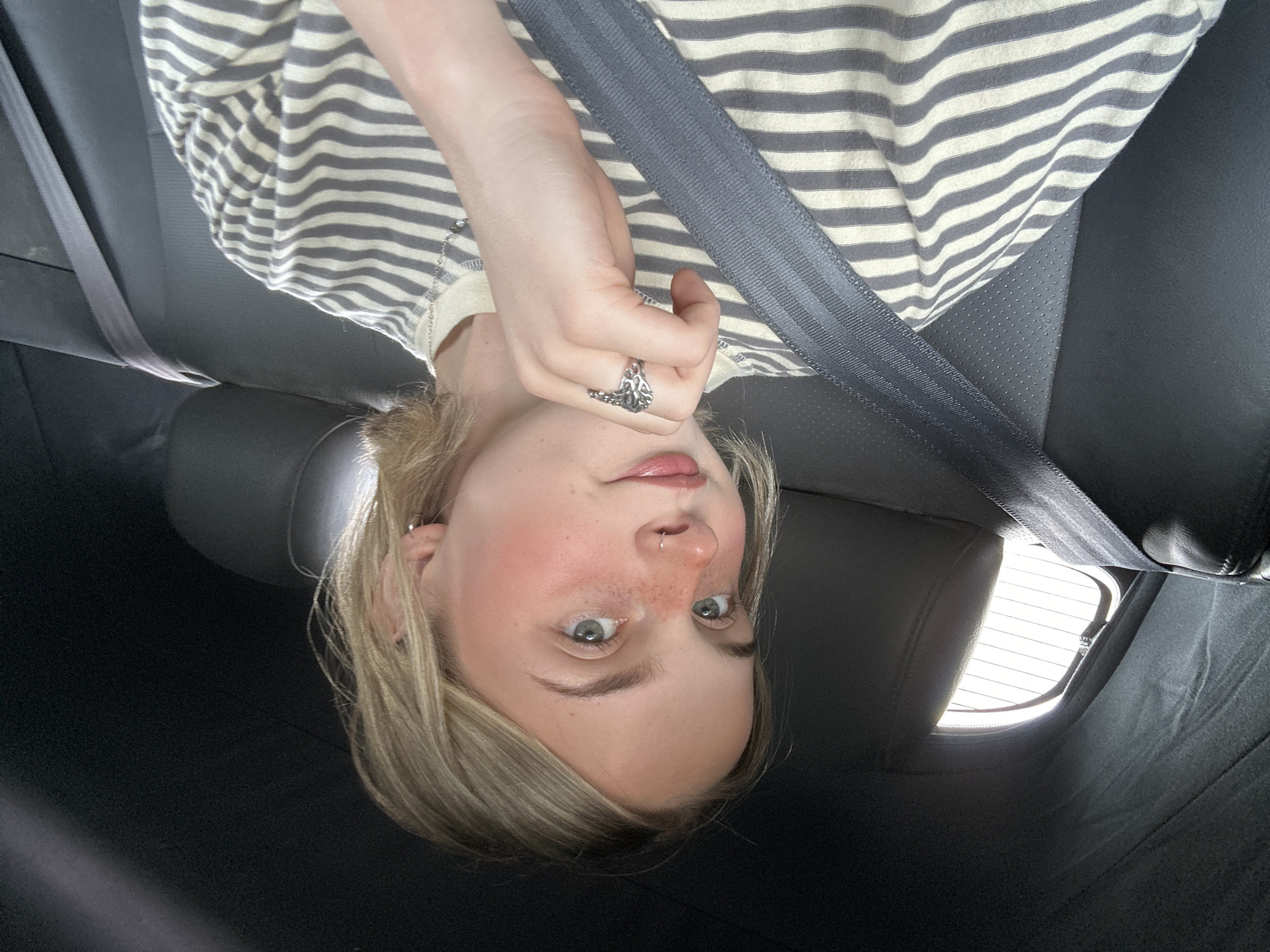 A young person with fair skin and light blonde hair is in the back seat of a vehicle, secured with a grey seatbelt. They are wearing a striped shirt and a ring on their finger. A portion of the vehicle's rear window and interior are visible. The location in Kelowna, Canada is not discernible from the image.